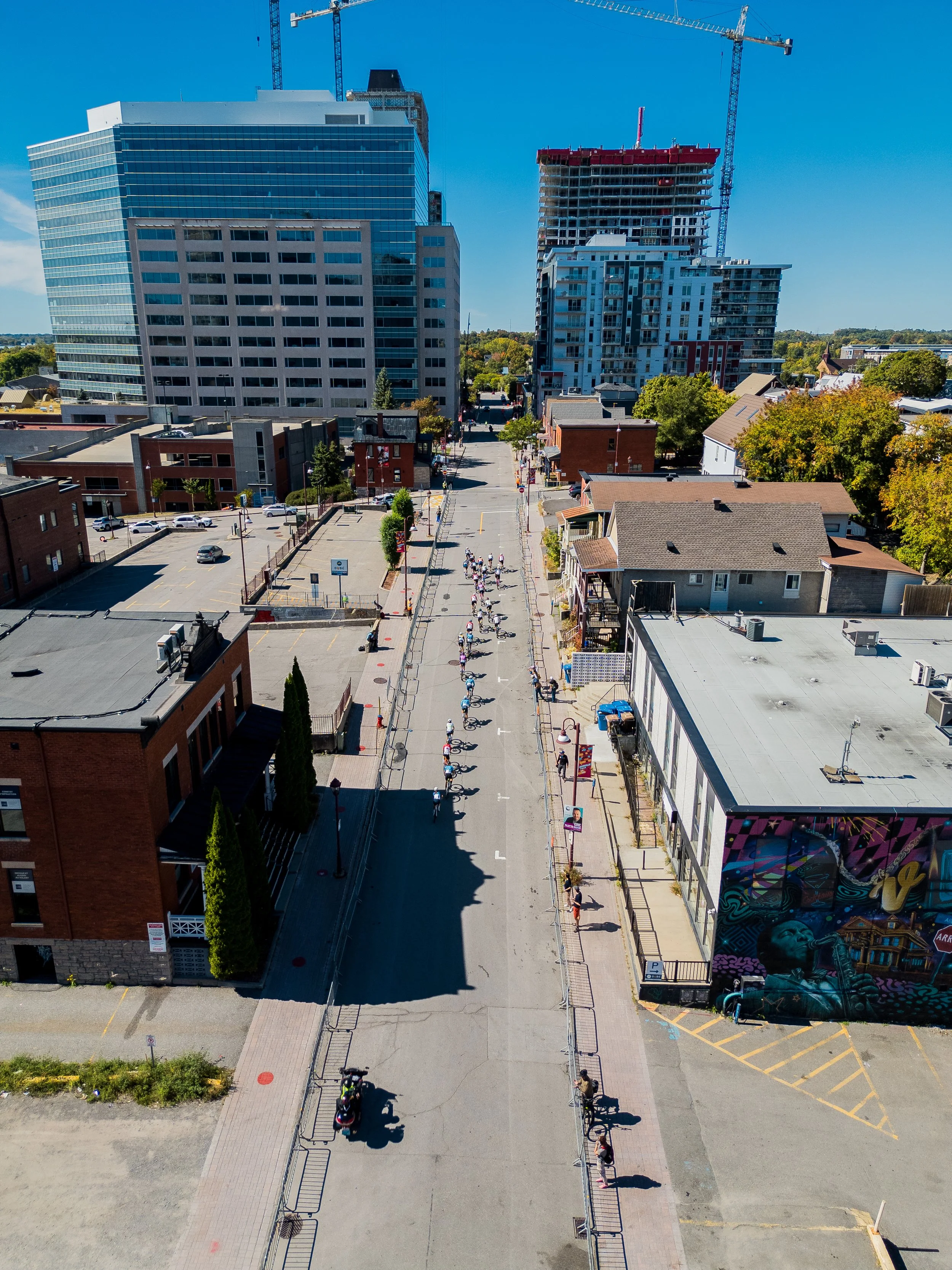 Aerial view of a city street lined with buildings, including a large modern office building, a high-rise under construction with cranes, and smaller storefronts with murals, with pedestrians and cyclists on a sunny day.