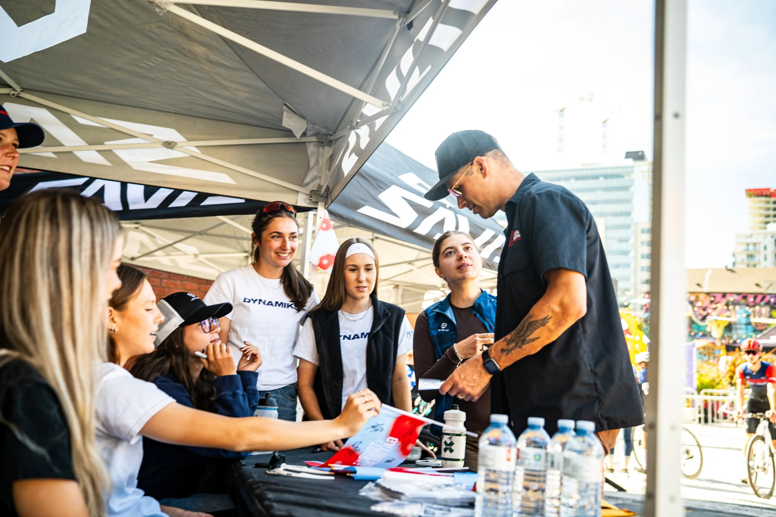 Group of young women signing autographs and getting photos at an outdoor event, standing under a tent with a cityscape in the background.