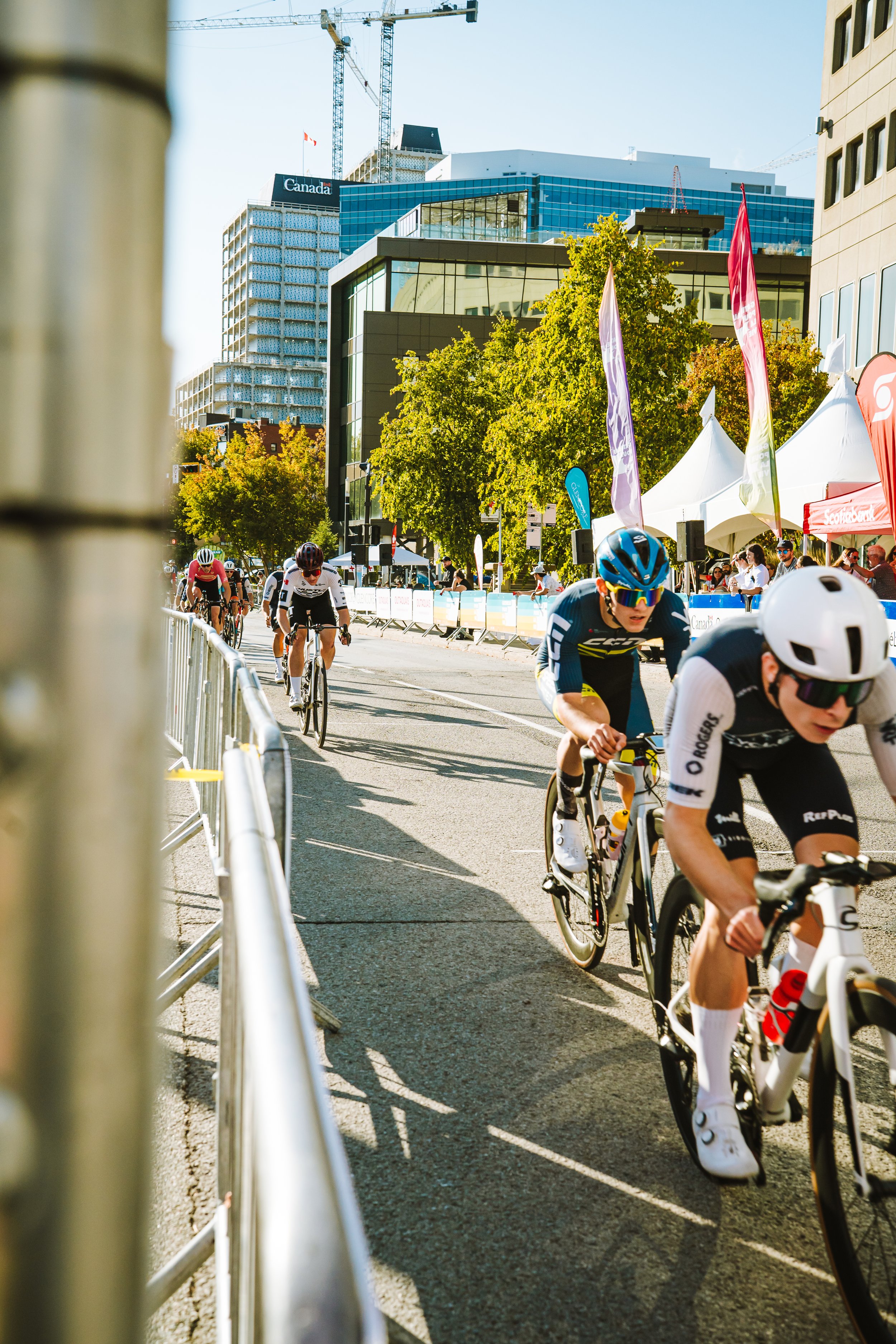 Cyclists competing in a race on a city street lined with barriers, trees, and modern buildings with construction cranes in the background.