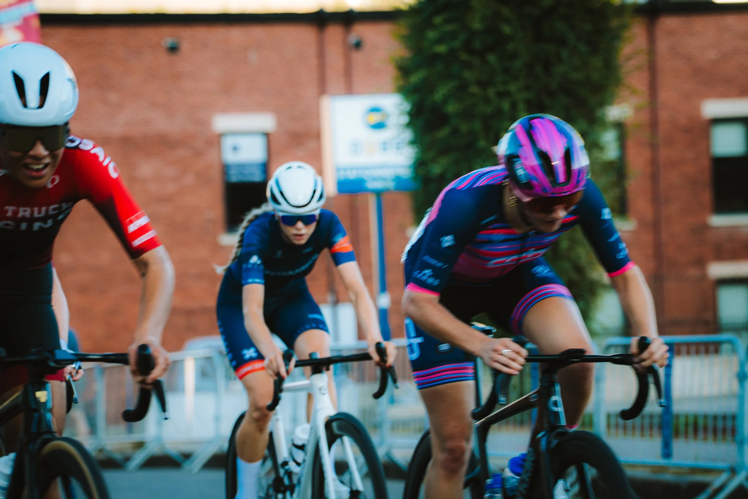 Cyclists racing on the street in a competitive cycling event, wearing helmets and sport gear, with brick buildings and trees in the background.