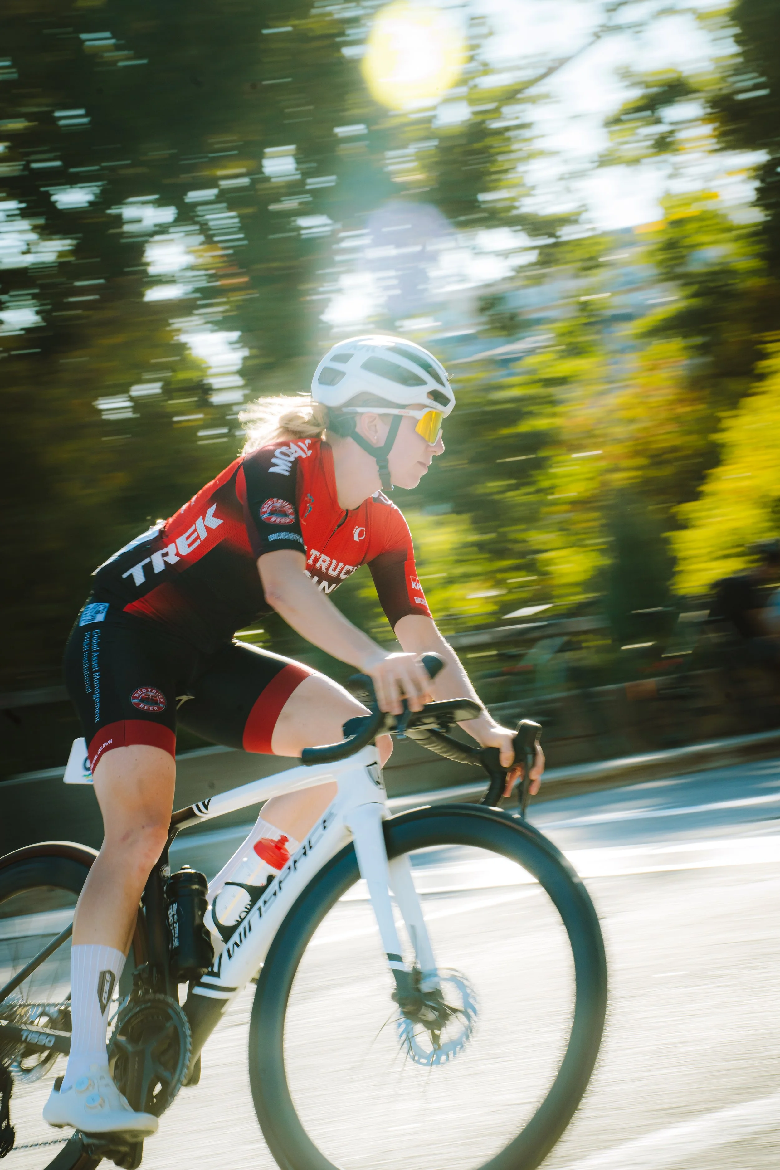 A female cyclist in a red and black jersey riding a white bike on a sunny day with blurred green trees in the background.