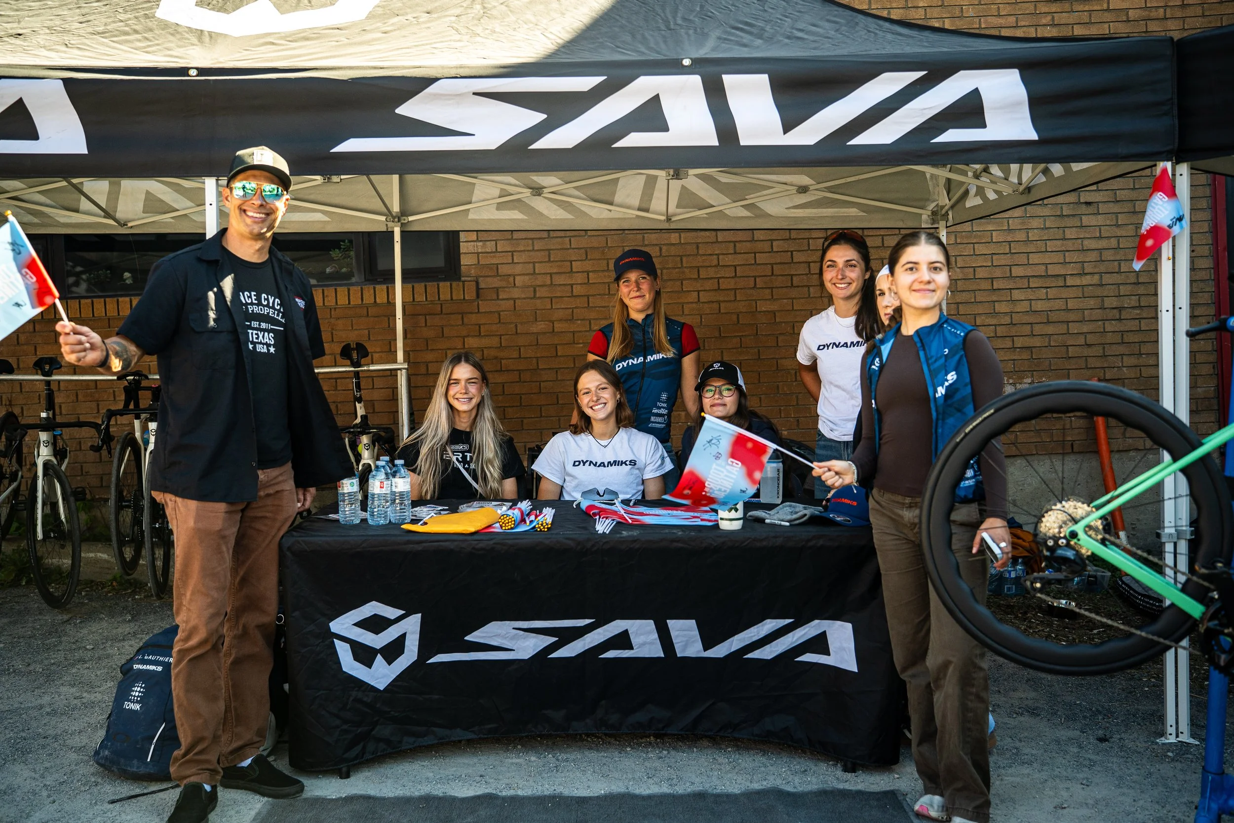 Six people smiling at an outdoor booth with a black tablecloth and a canopy. The booth has cycling-related items, and some people are holding small flags. Bicycles are visible in the background.