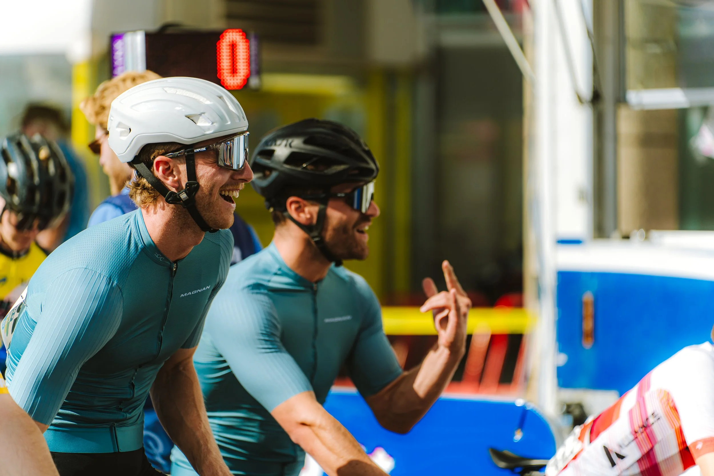 Two male cyclists wearing helmets and sunglasses laughing and talking at a cyclist event.