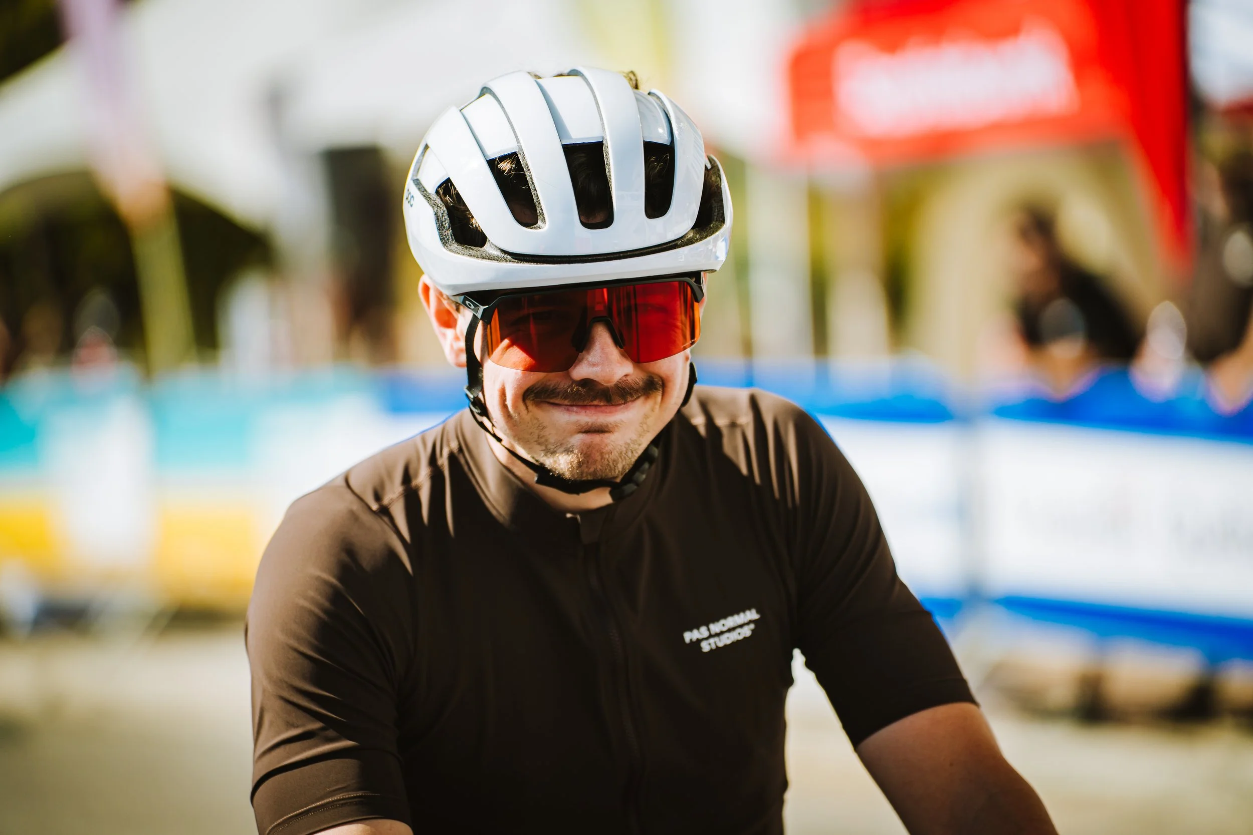 A man wearing a white bicycle helmet, red sports sunglasses, and a black cycling jersey smiling outdoors with blurry colorful banners in the background.