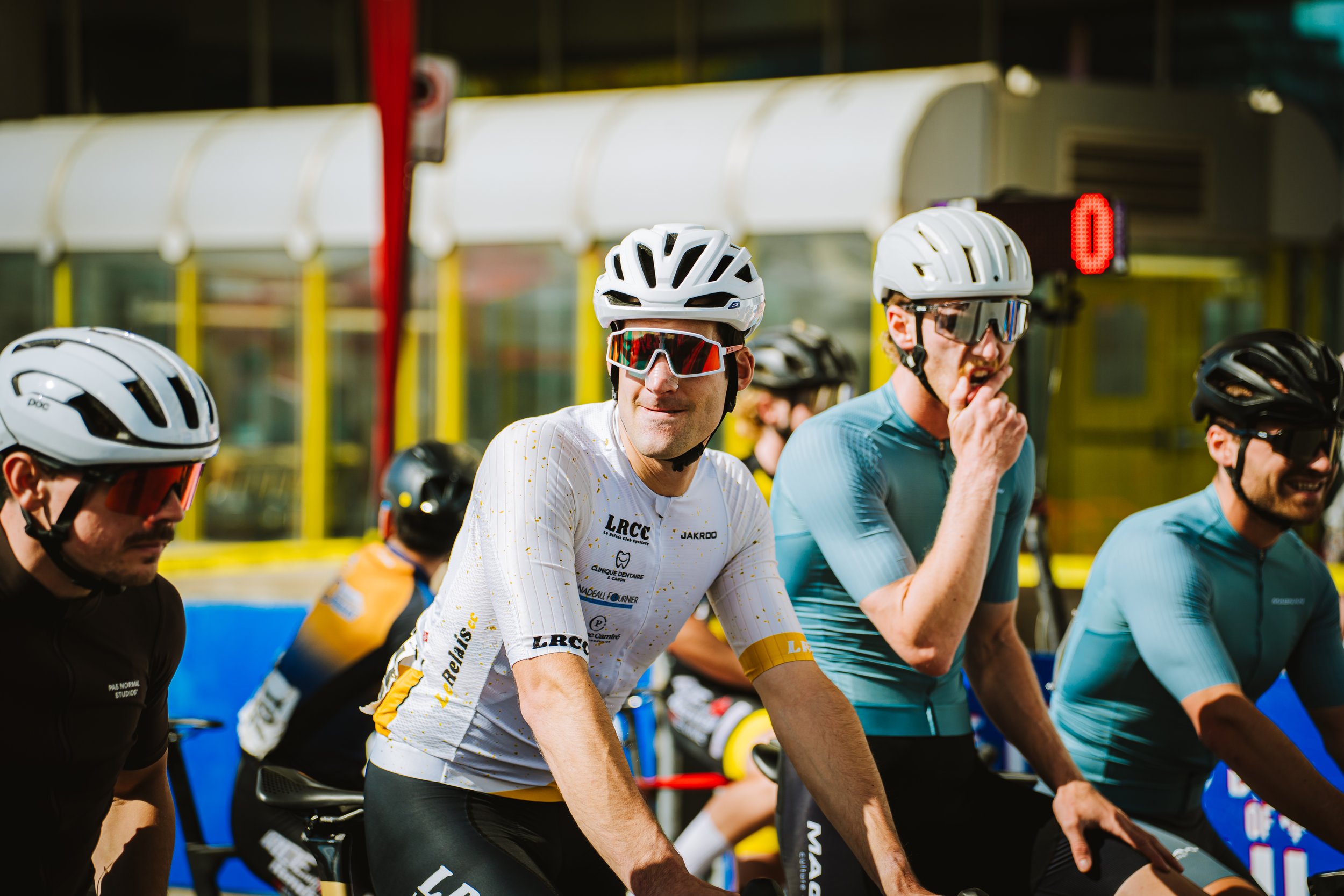 Cyclists wearing helmets and sunglasses, preparing for a race or ride, with a busy urban background.