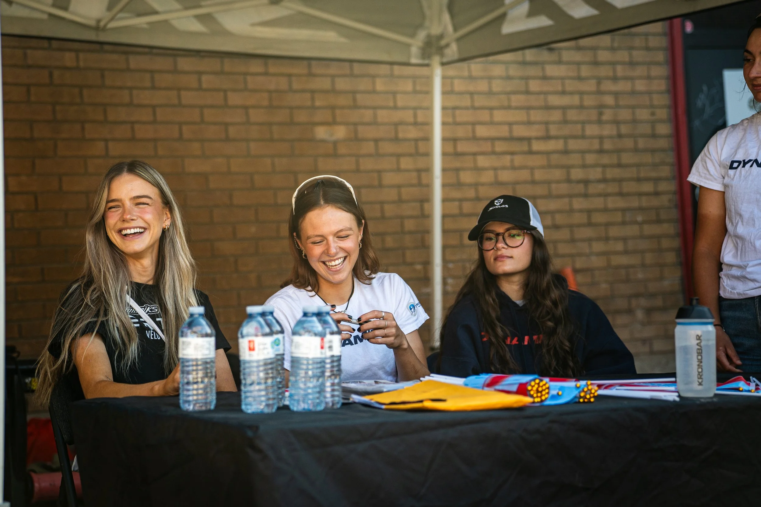 Three women sitting at a table under a canopy, laughing and smiling, with water bottles and colorful lanyards or ribbons on the table.
