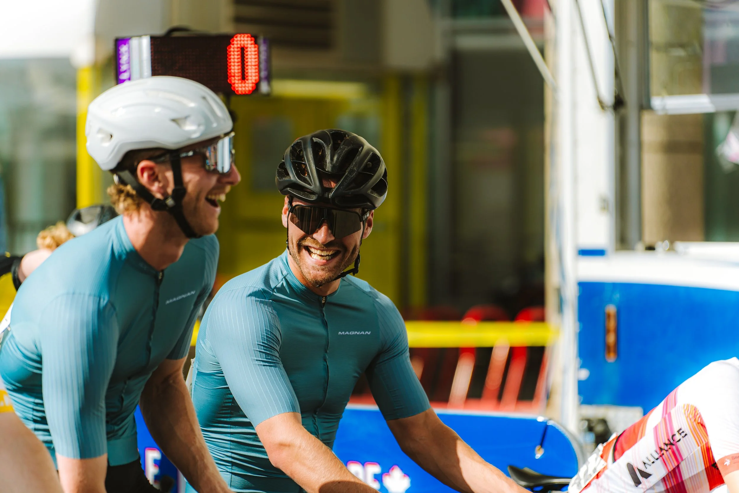 Two male cyclists wearing blue jerseys, helmets, and sunglasses, laughing and smiling together after a race, with a yellow barrier and a digital clock in the background.