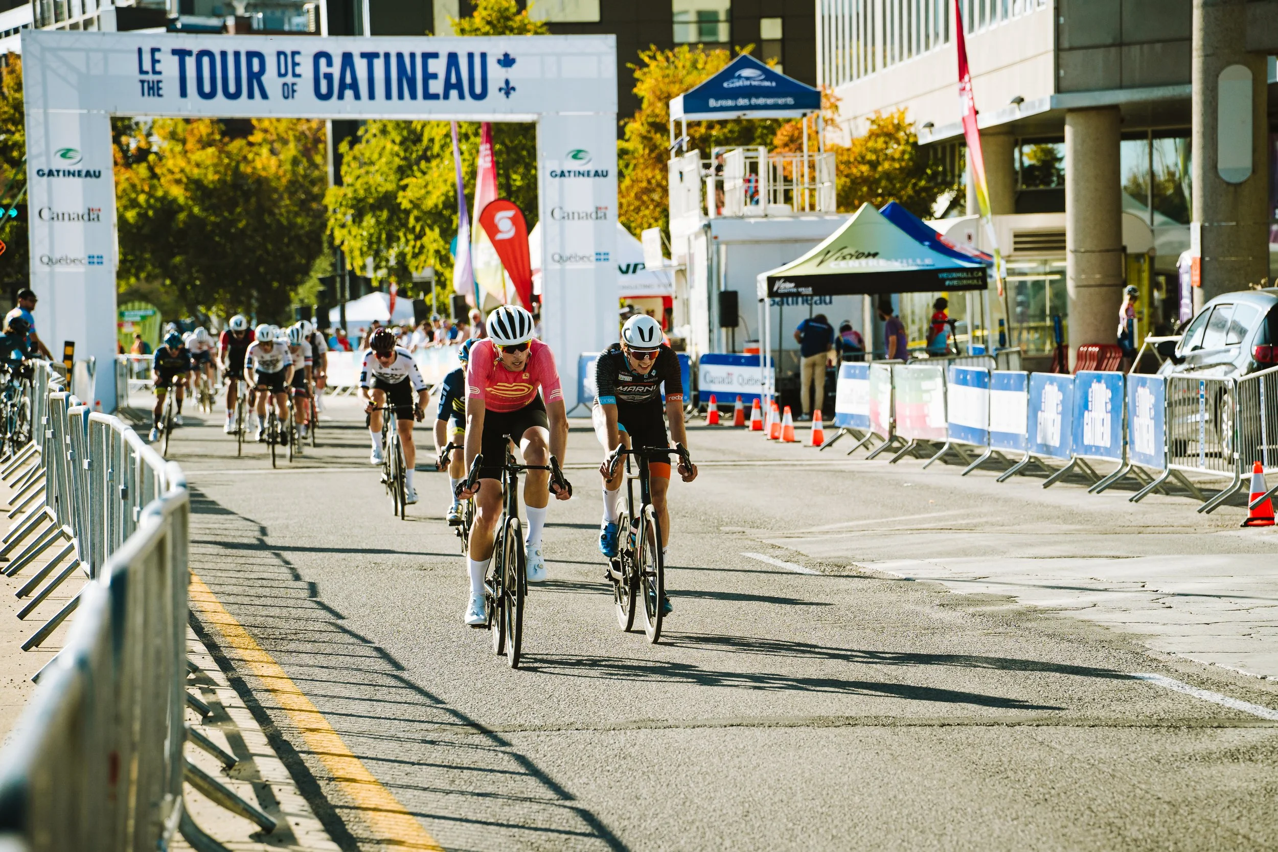 Cyclists riding in a race under a large archway with the text "Le Tour de Gatineau" during daytime. There are barriers and tents along the street, with trees and a building in the background.