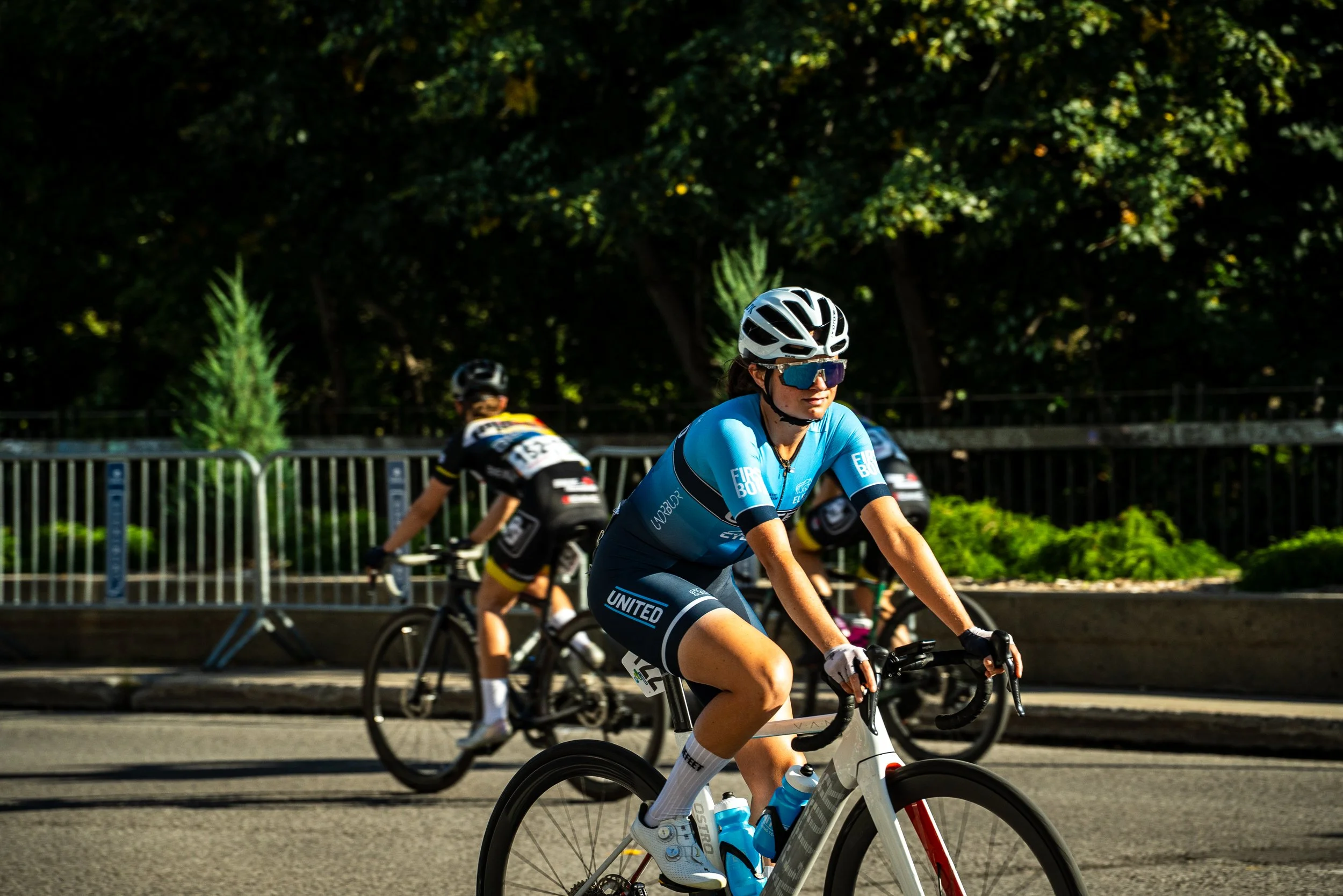 Cyclist in blue gear and white helmet riding a bike during a race, with other cyclists and green trees in the background.