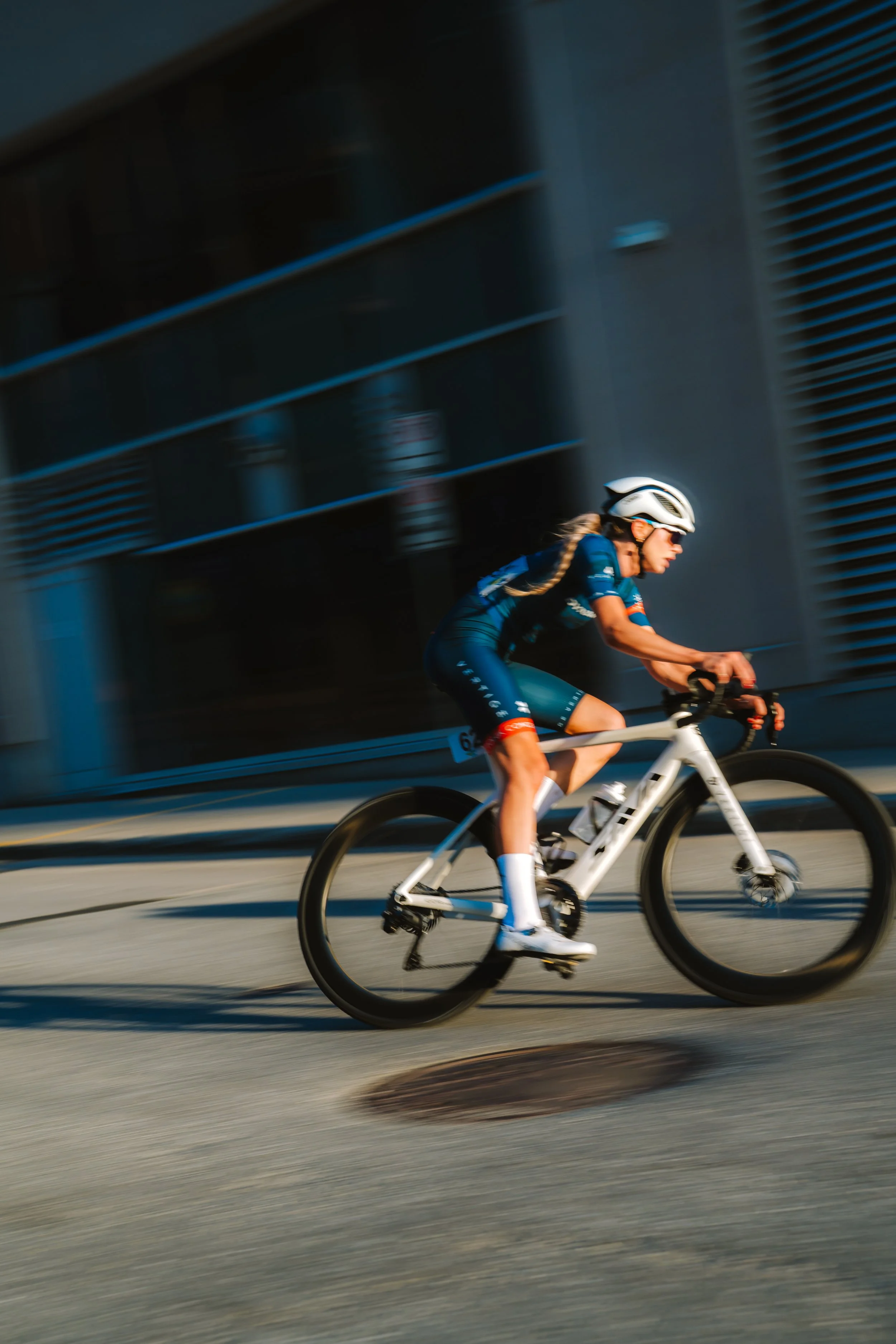 A woman in a blue cycling outfit and white helmet riding a bike on an urban street, with motion blur indicating high speed.