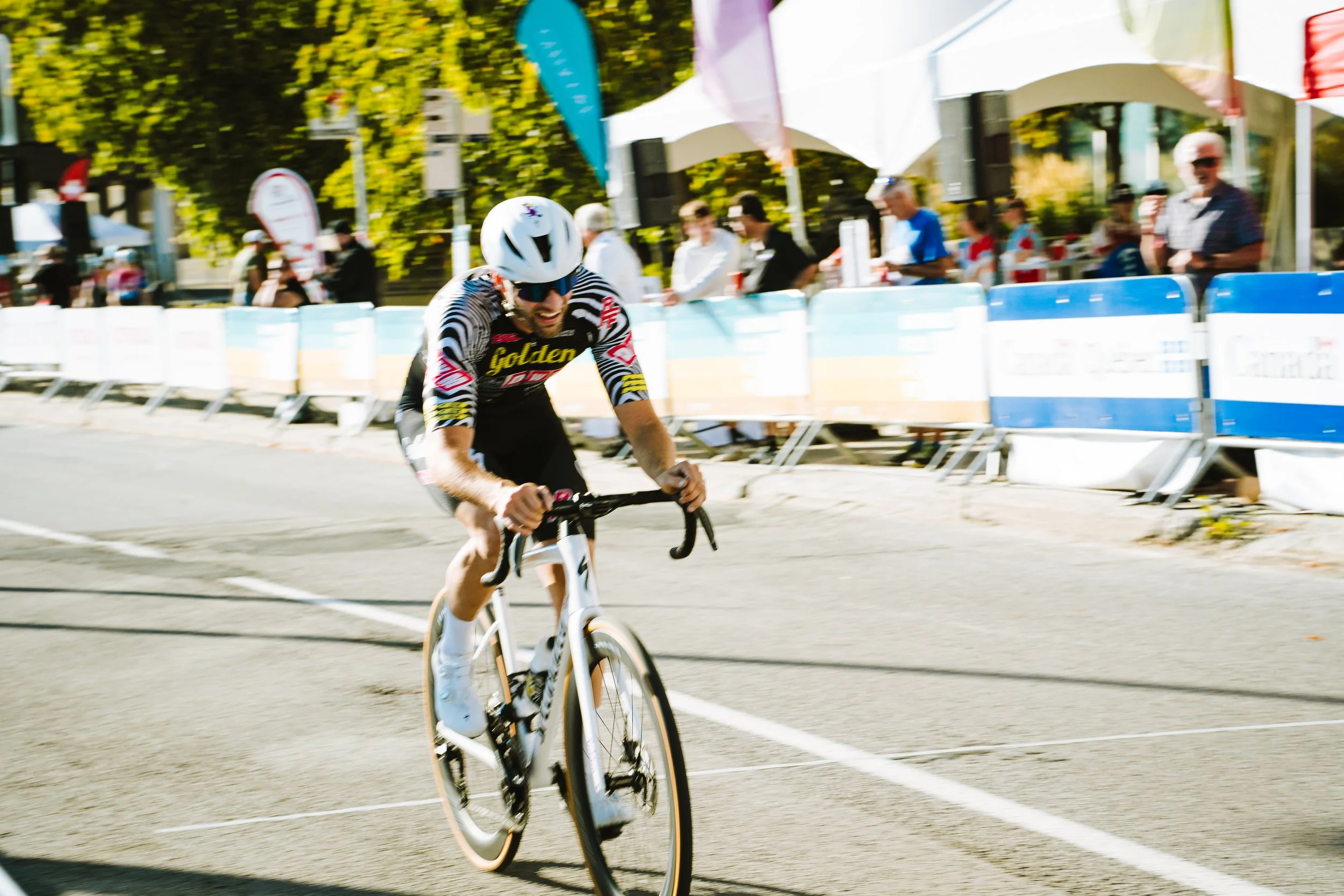 Cyclist in a black and white striped jersey with yellow and red accents, wearing a white helmet, racing on a street with spectators standing behind barricades under tents.