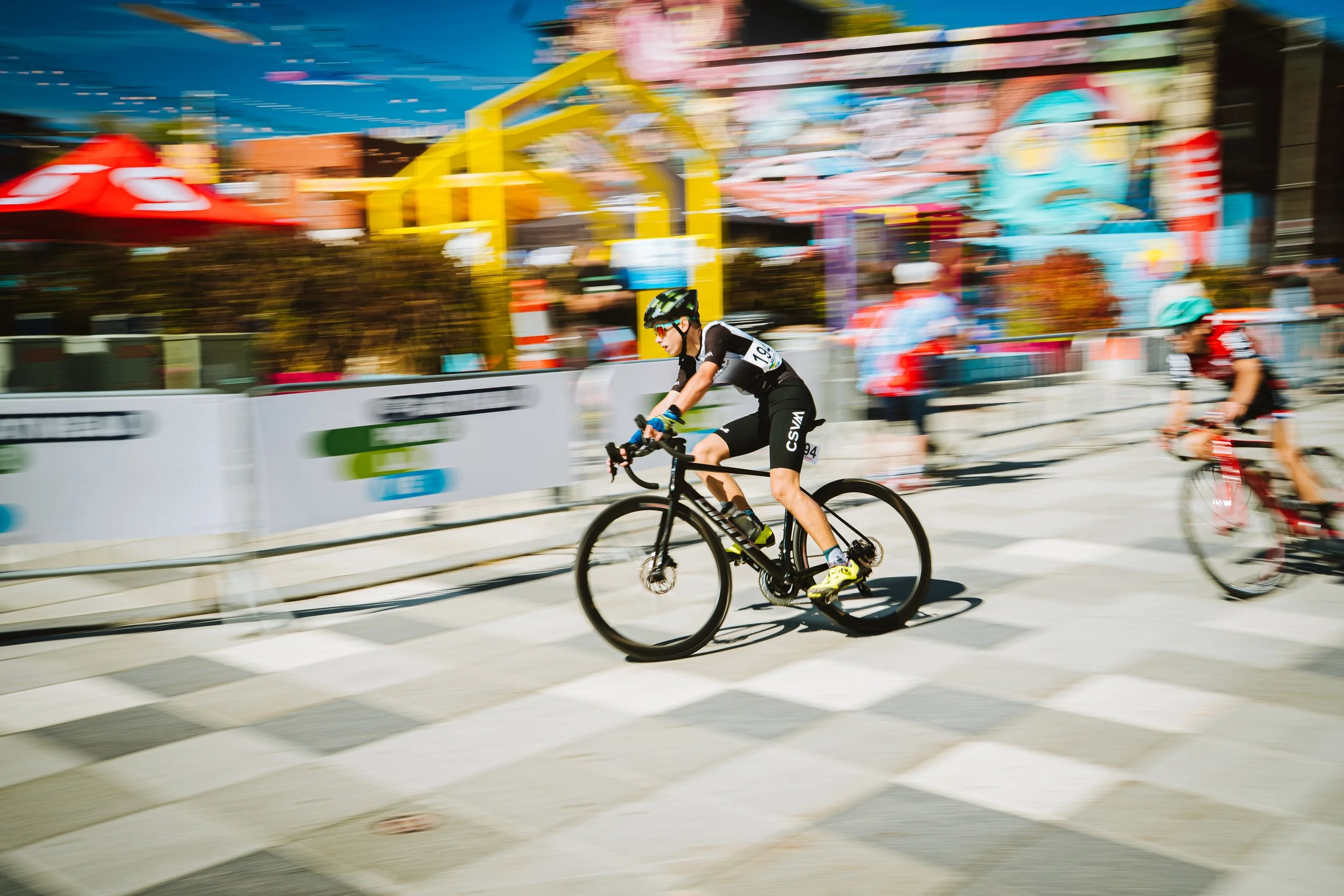 A cyclist riding a black bicycle during a race, with motion blur showing speed, in an outdoor setting with colorful amusement park attractions in the background.