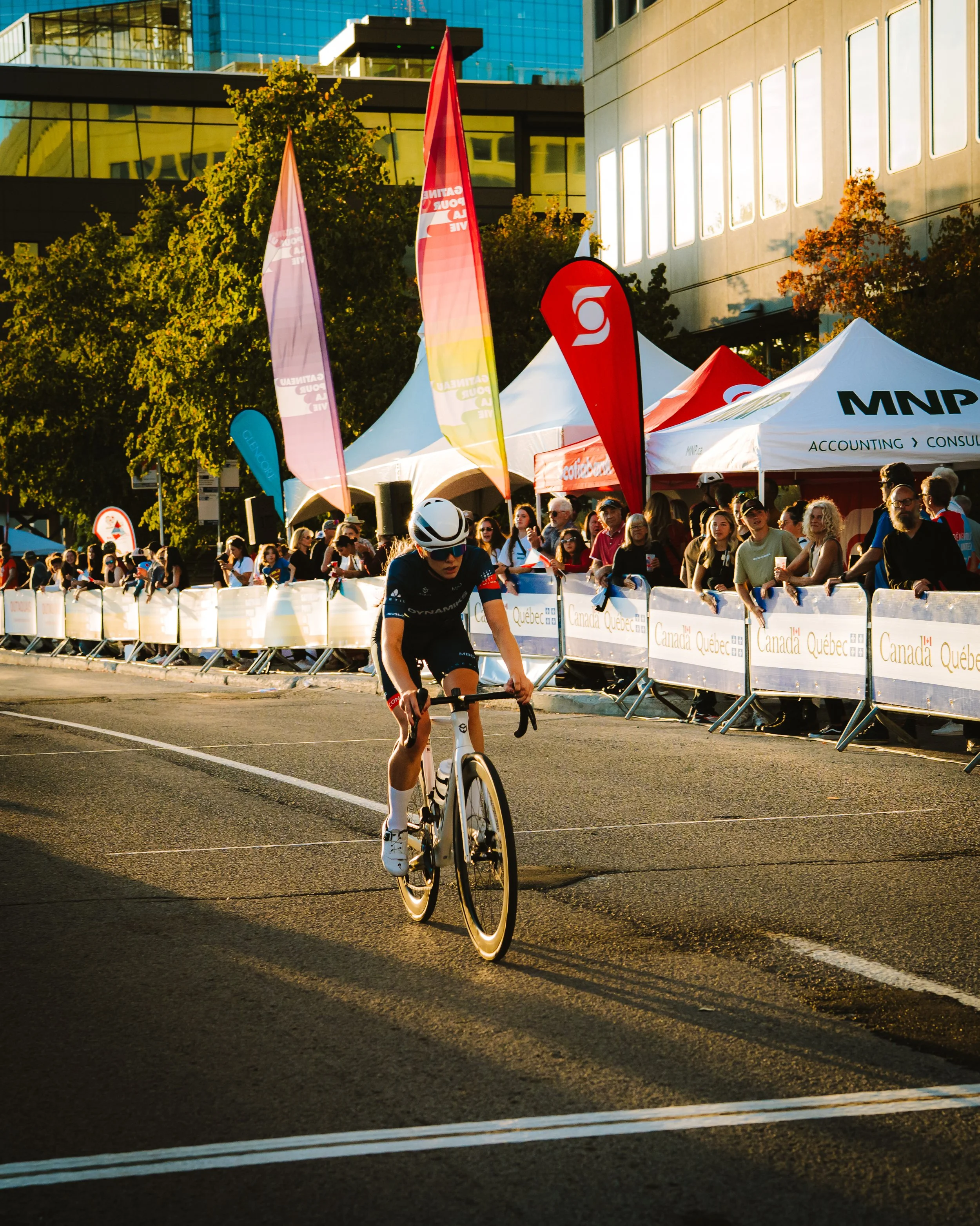 A cyclist riding on a city street during a race with spectators behind barriers and tents along the sidewalk, flags, and banners in the background.