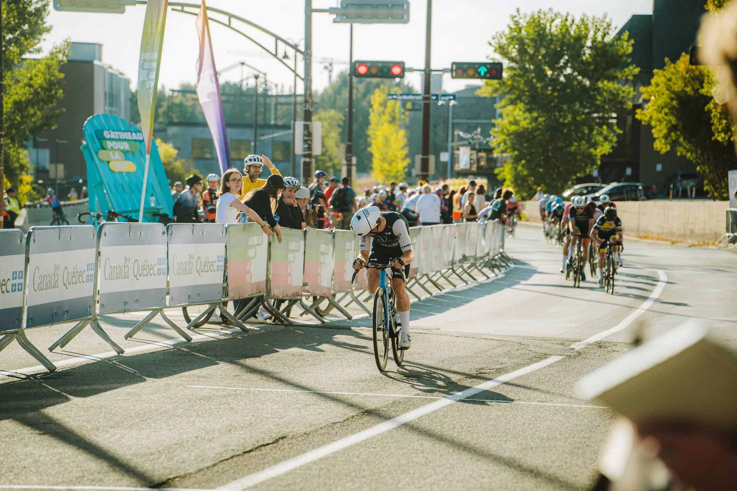 Cyclist finishing a race while supporters cheer from behind barriers labeled 'Canada Quebec' during a sunny day race in an urban area.