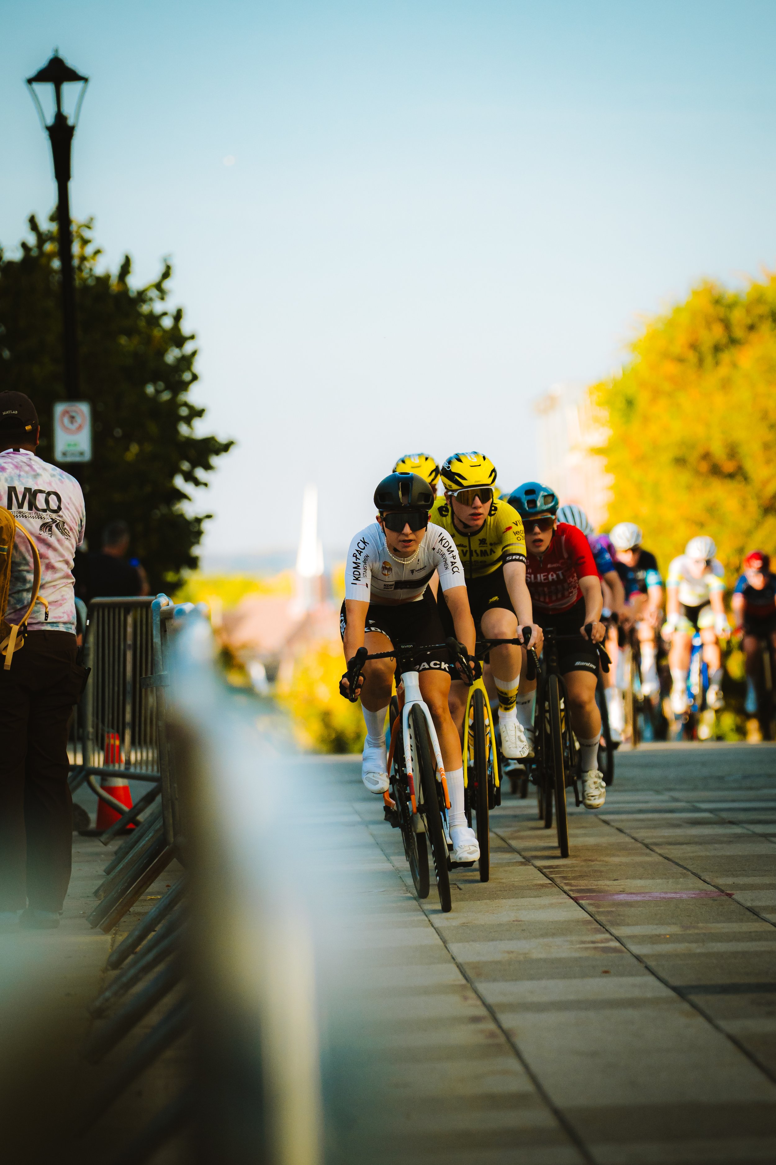 Group of cyclists racing outdoors on a paved path, with a crowd watching on the side, trees with fall foliage, and a clear sky.
