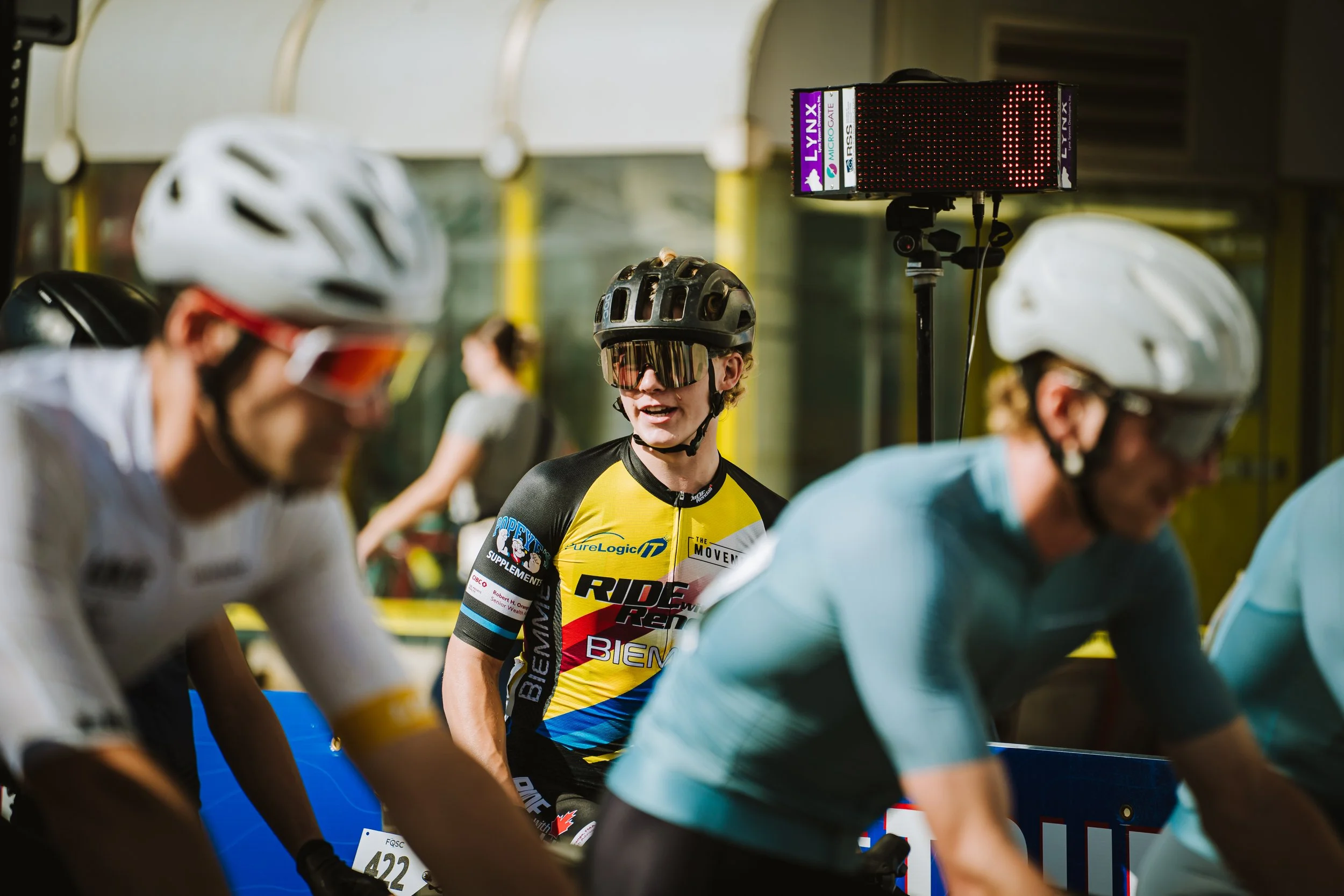 Cyclists preparing at the start line of a race, with one cyclist in focus wearing a colorful jersey and helmet.