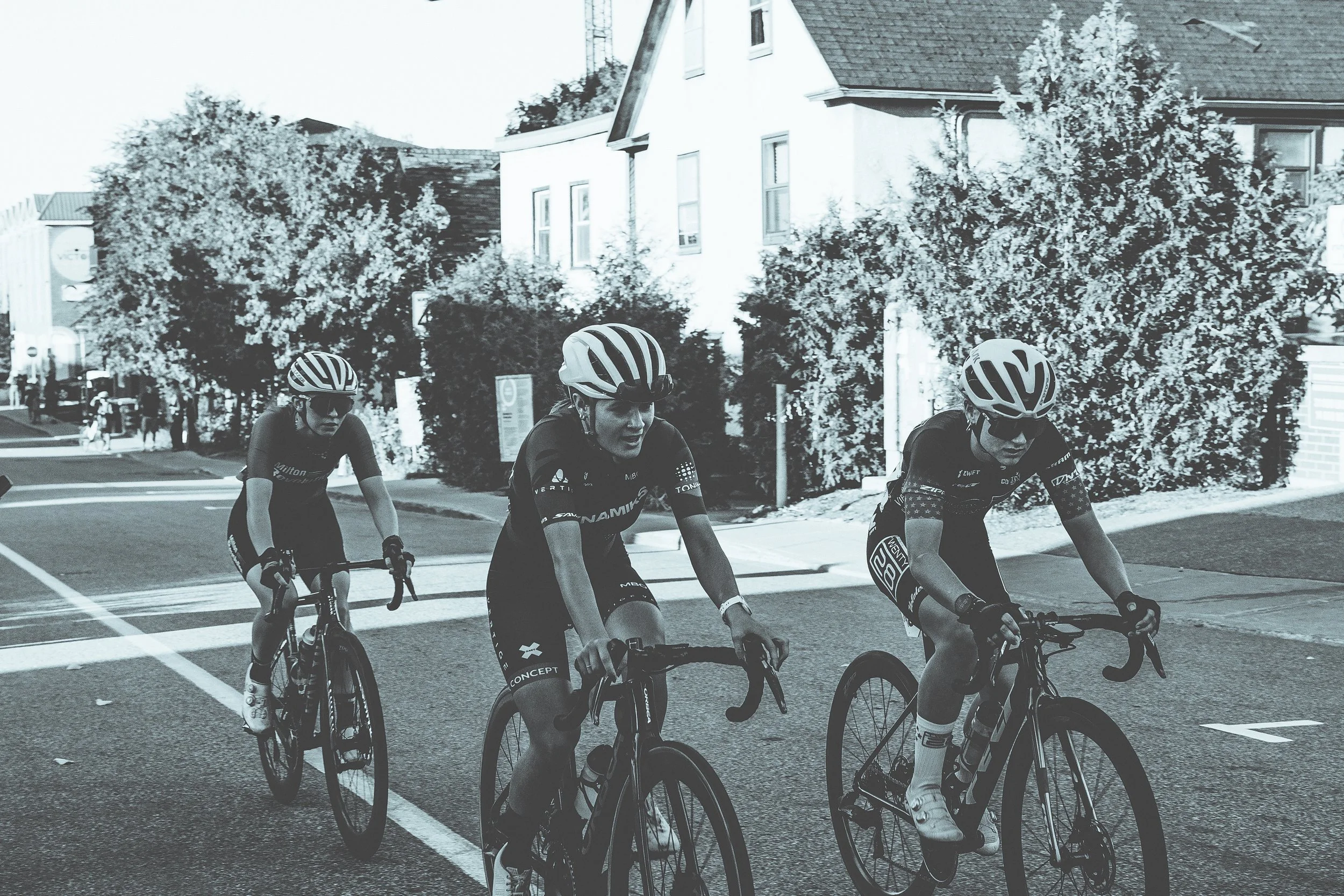 Three cyclists riding on a city street, wearing helmets and sports gear, with trees and residential buildings in the background.