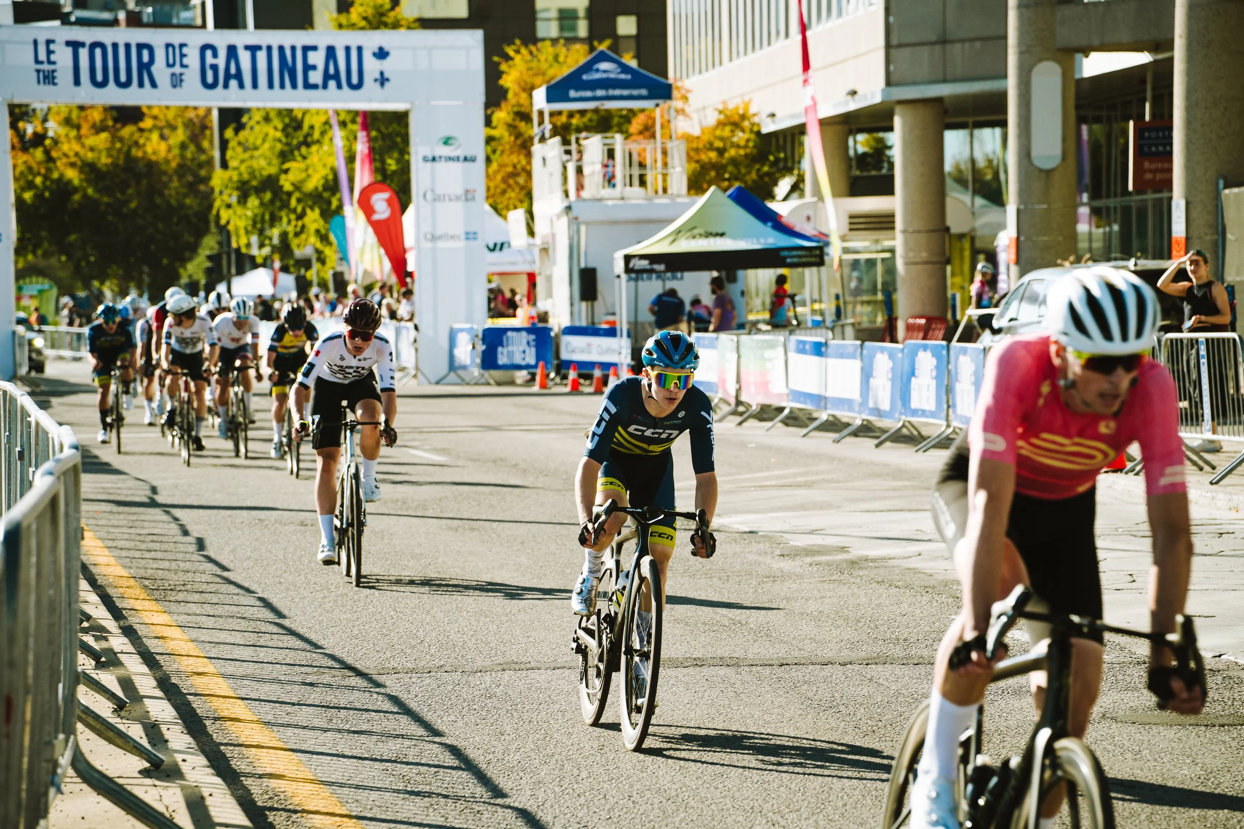 Cyclists competing in a race called 'Le Tour de Gatineau' on a city street with spectators and race infrastructure around.