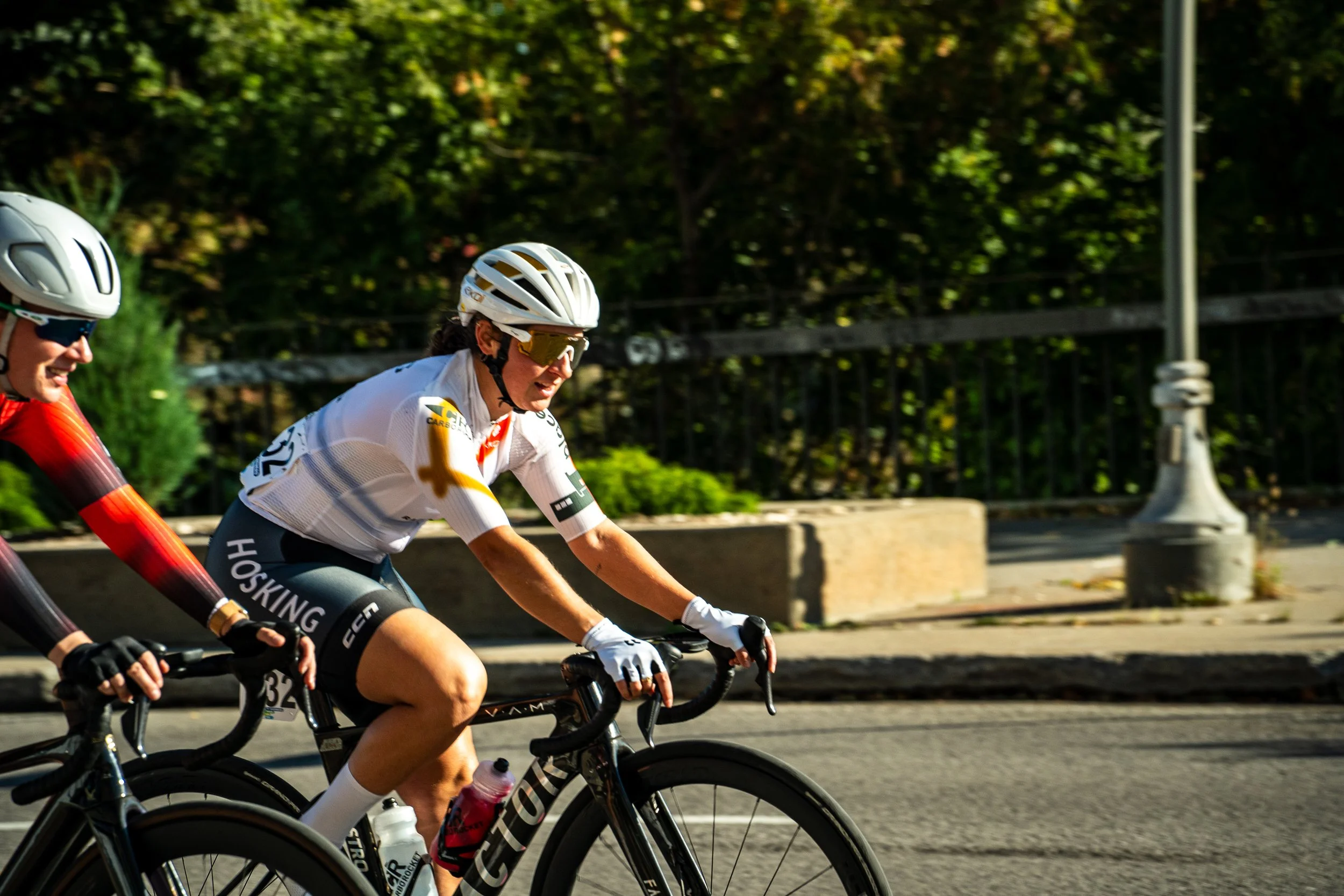 Two female cyclists racing on a paved road, wearing helmets and sunglasses, with a background of trees and a sidewalk.