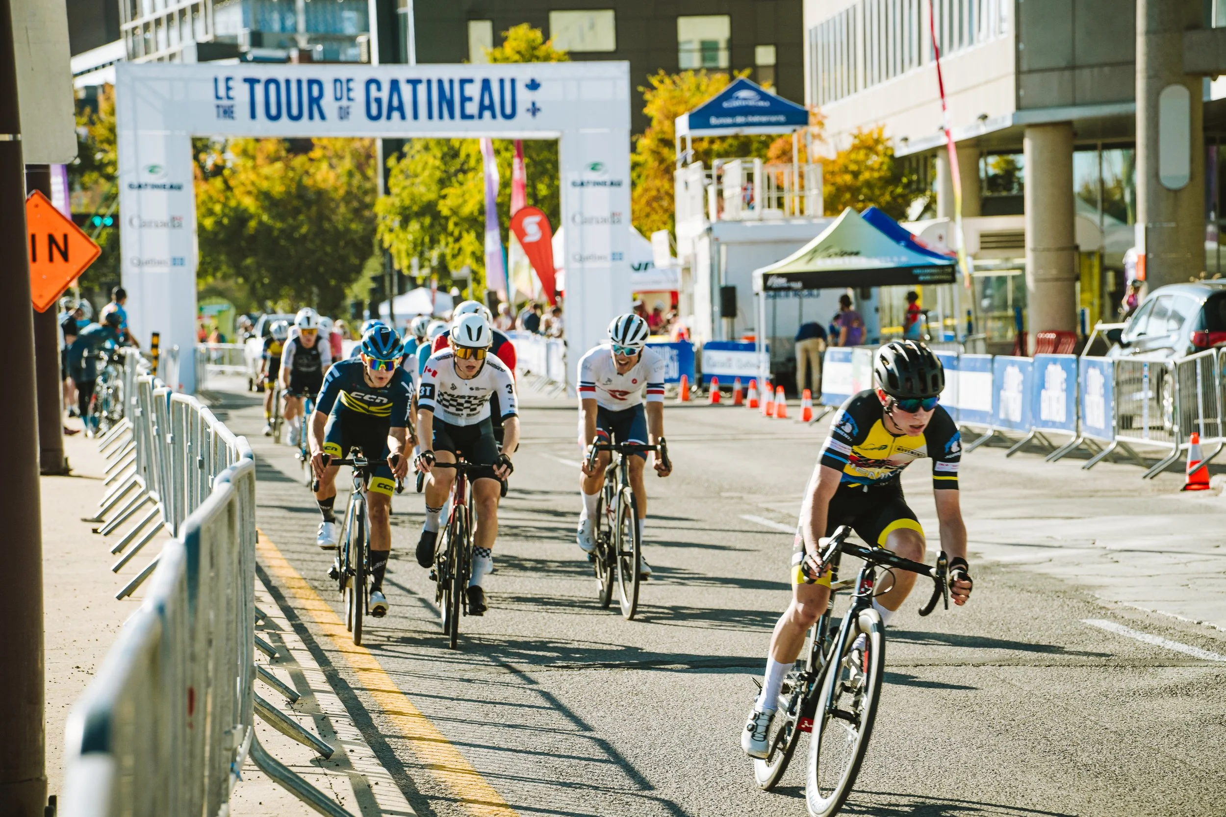 Cyclists competing in the Tour de Gatineau race, riding on city streets with spectators and event banners in the background.