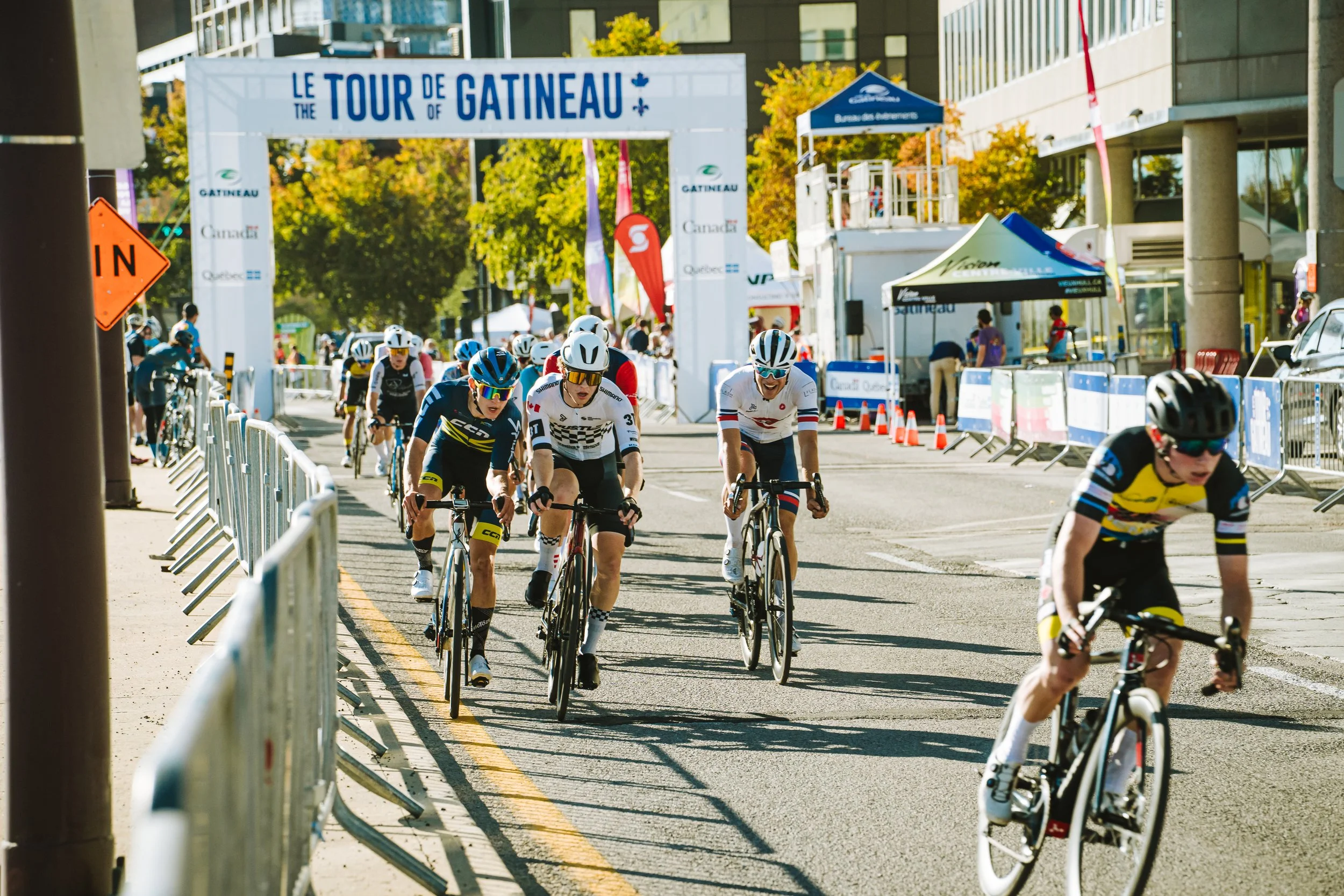 Cyclists participating in a race on a city street during the Tour de Gatineau, with a start/finish arch and barricades lining the route.