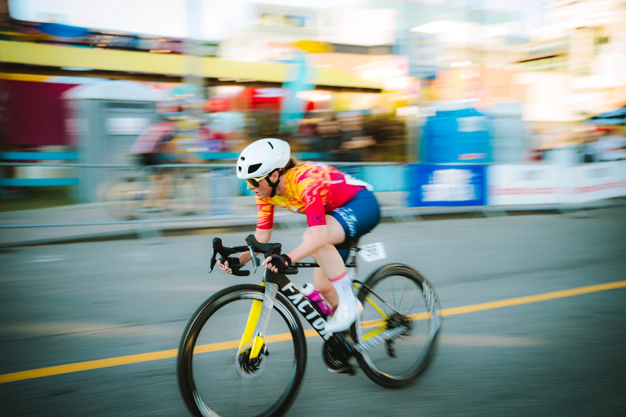 A cyclist wearing a white helmet, sunglasses, a colorful red and yellow jersey, black shorts, and white shoes rides a yellow and black bicycle on a city street during a race, with motion blur in the background.