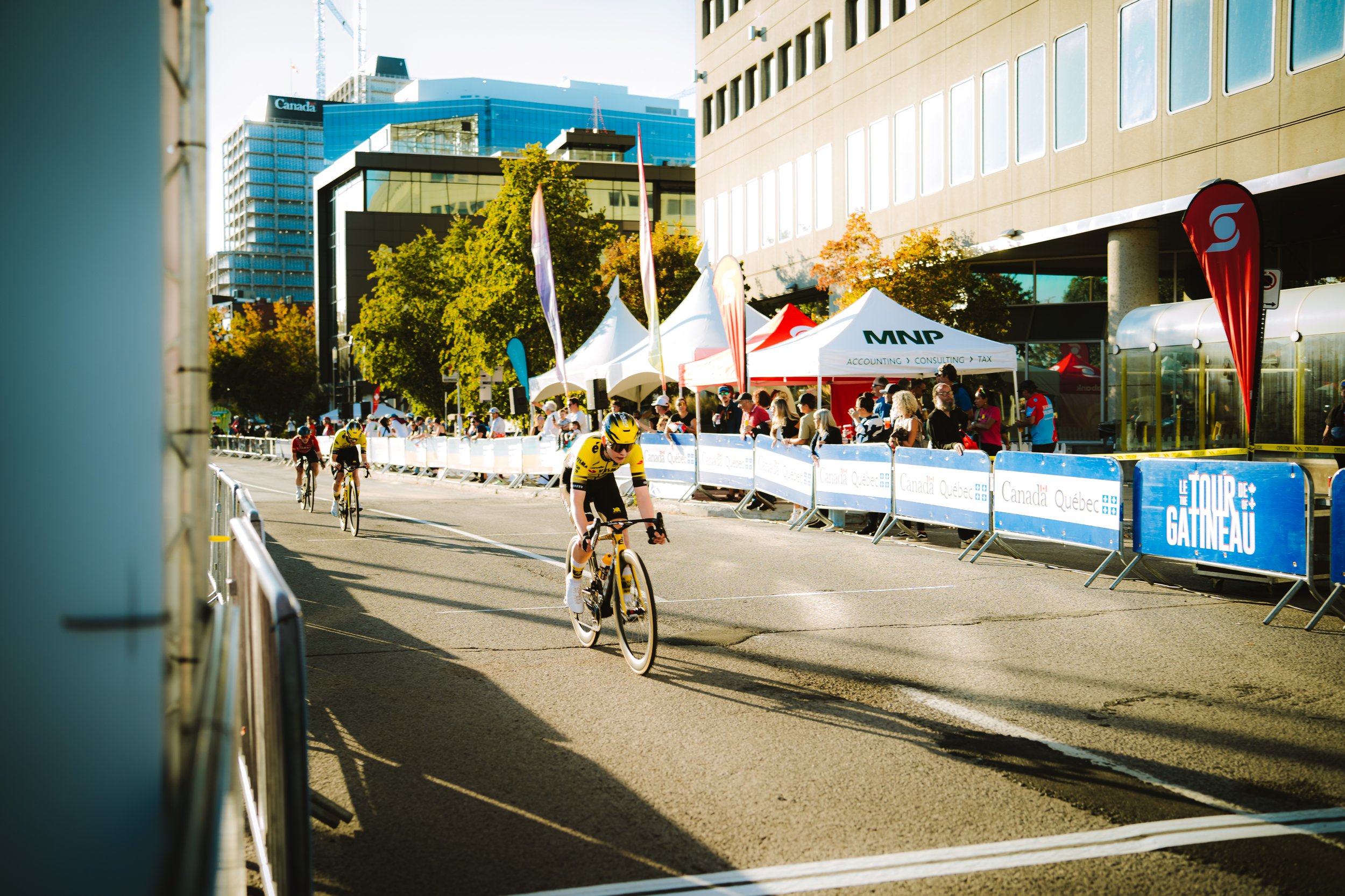Cyclists racing on a city street during a cycling event, with spectators and tents lining the sidewalk, under a clear sky with tall buildings in the background.