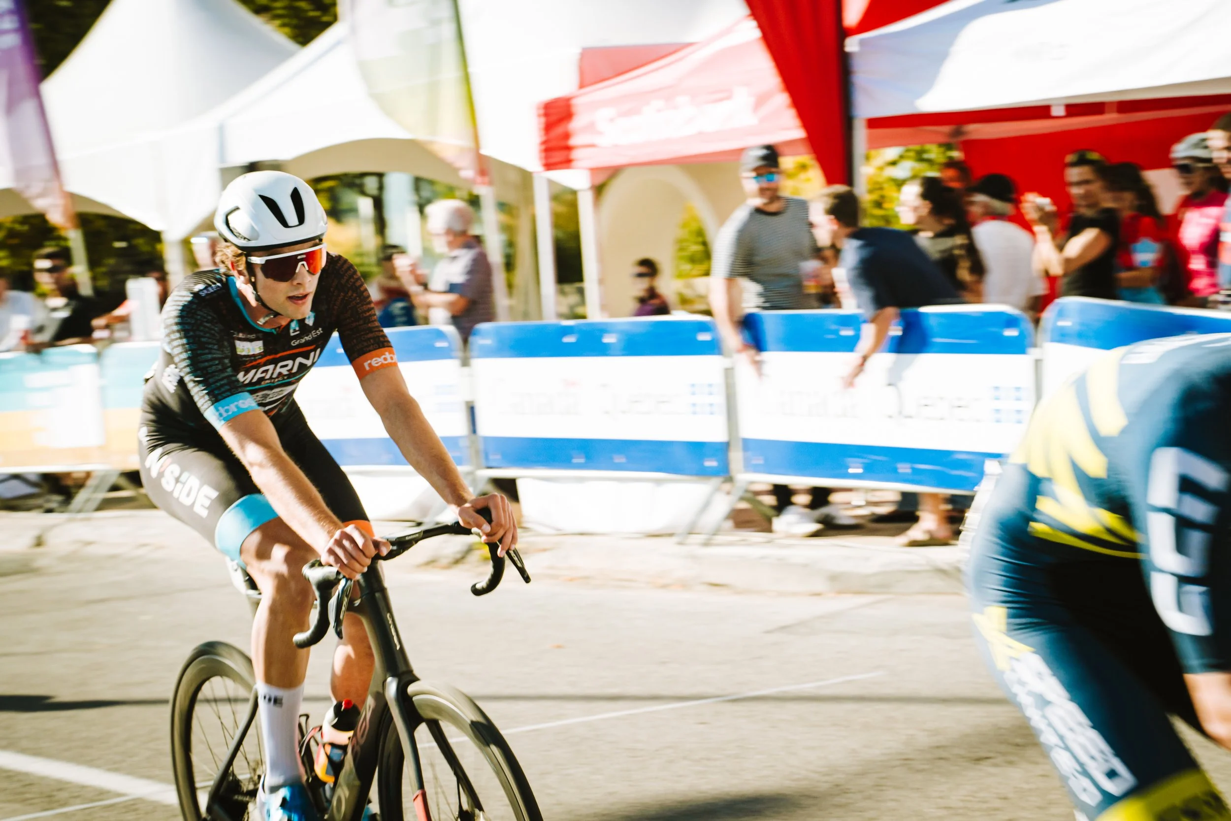 A cyclist wearing a helmet, sunglasses, and a black and blue cycling jersey competing in a race, with spectators and tents in the background.