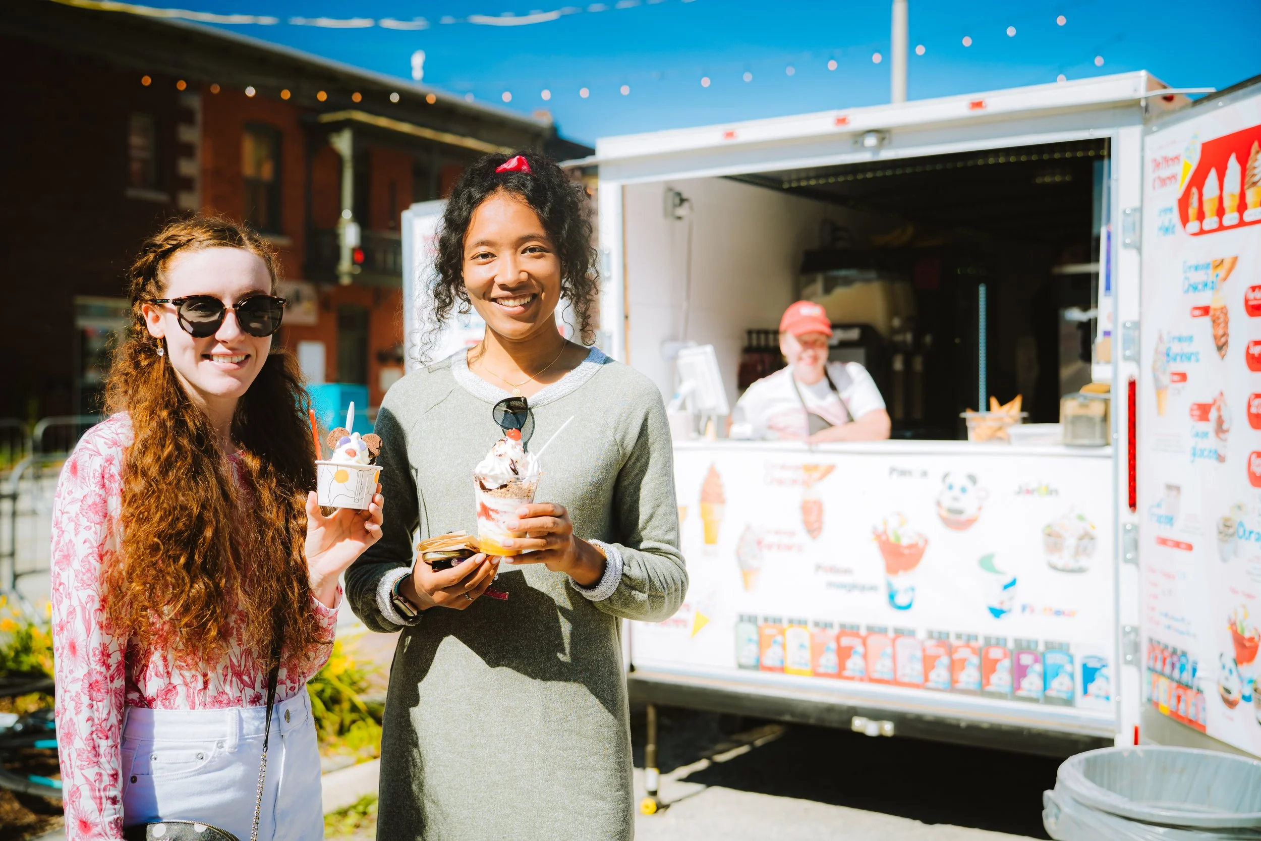 Two women smiling and holding ice cream cones at an outdoor ice cream stand, with a woman working behind the stand in the background.