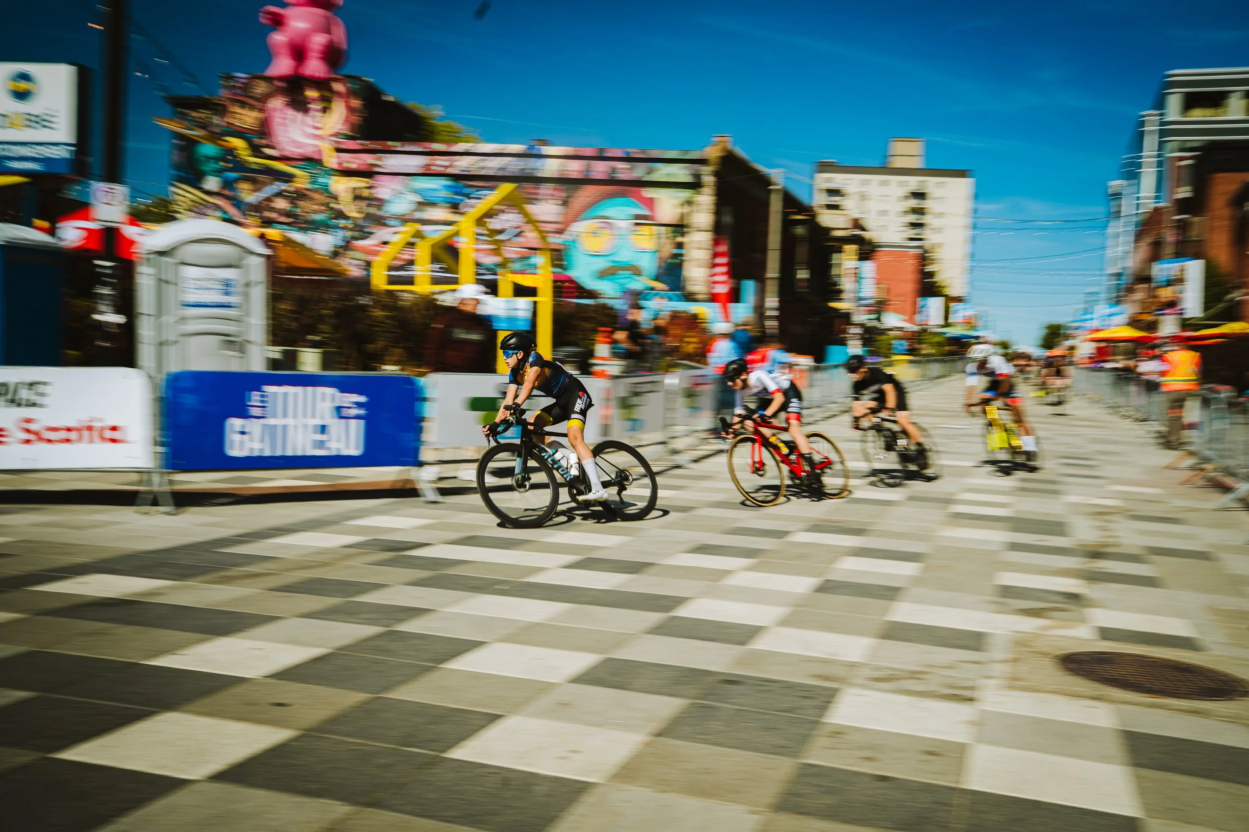 Cyclists participating in a bike race crossing a city street with checkered crosswalk, spectators on the sides, and colorful outdoor amusement park rides and murals in the background on a sunny day.