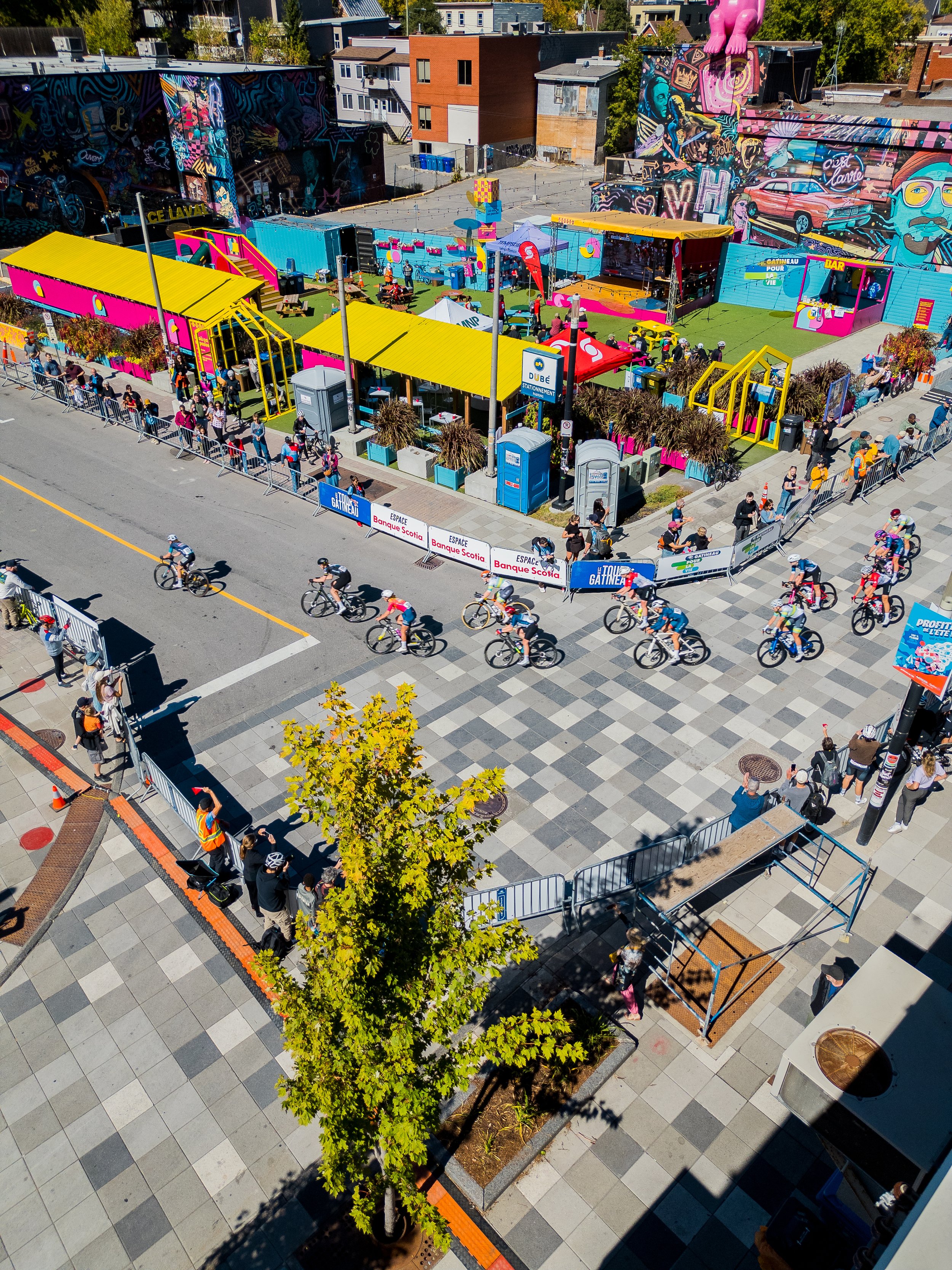 A colorful outdoor event with a cycling race on a city street, surrounded by spectators, barricades, and vibrant murals on nearby buildings.