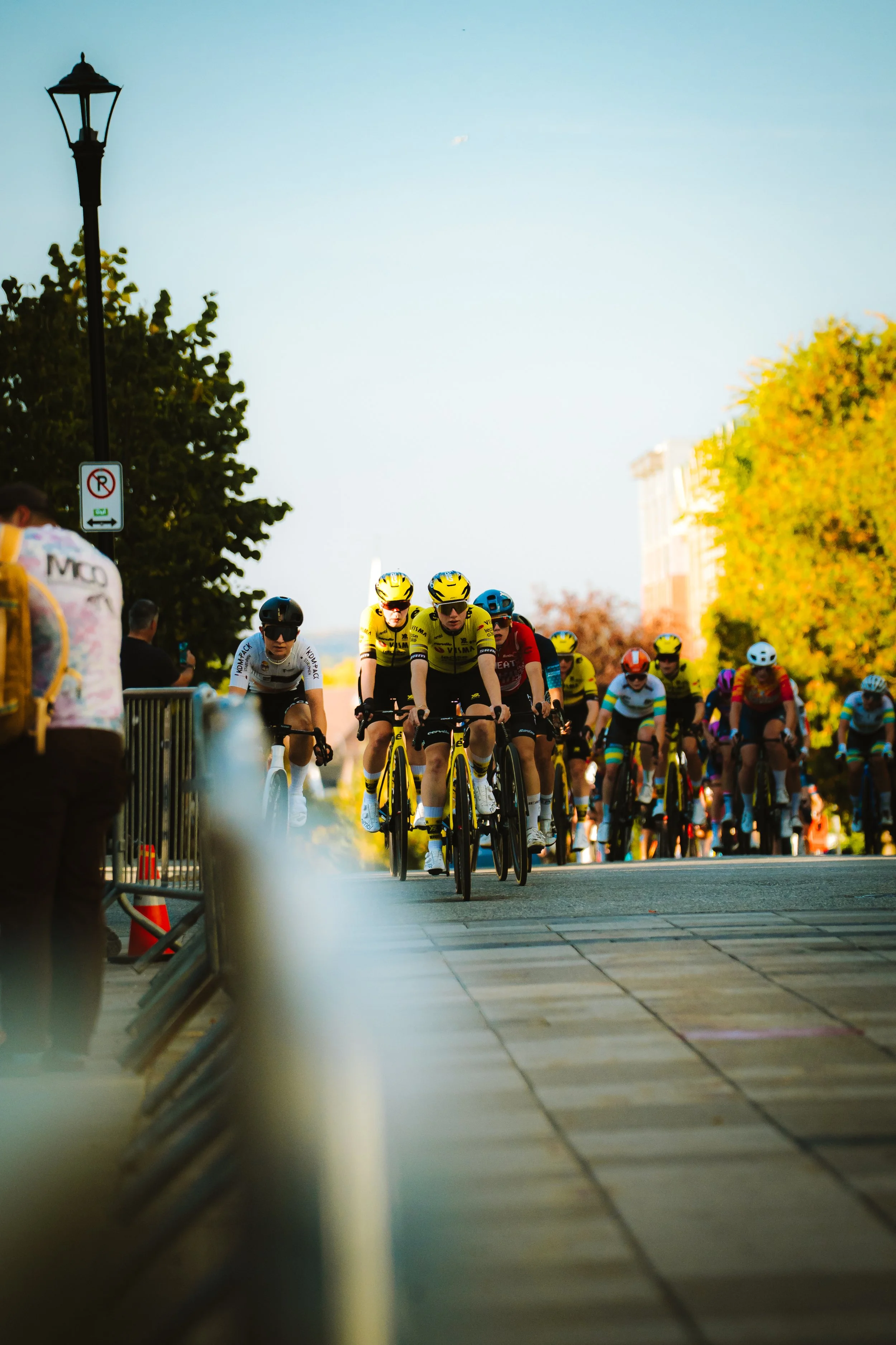 Group of cyclists participating in a race on a city street, with spectators along the sidewalk and trees with autumn foliage in the background.