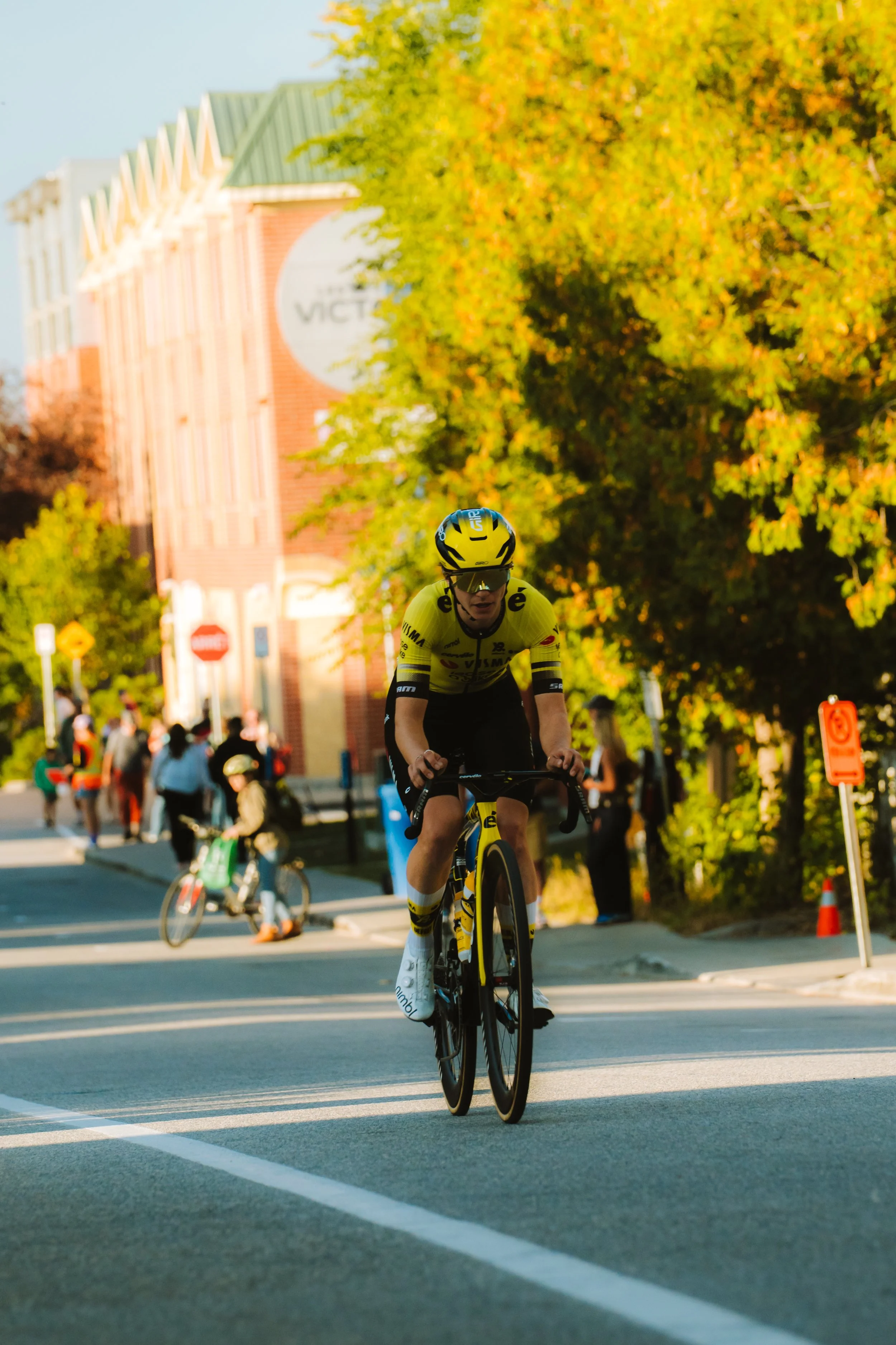 A cyclist wearing a yellow and black jersey and helmet riding a bike on a city street during daytime with trees and pedestrians in the background.