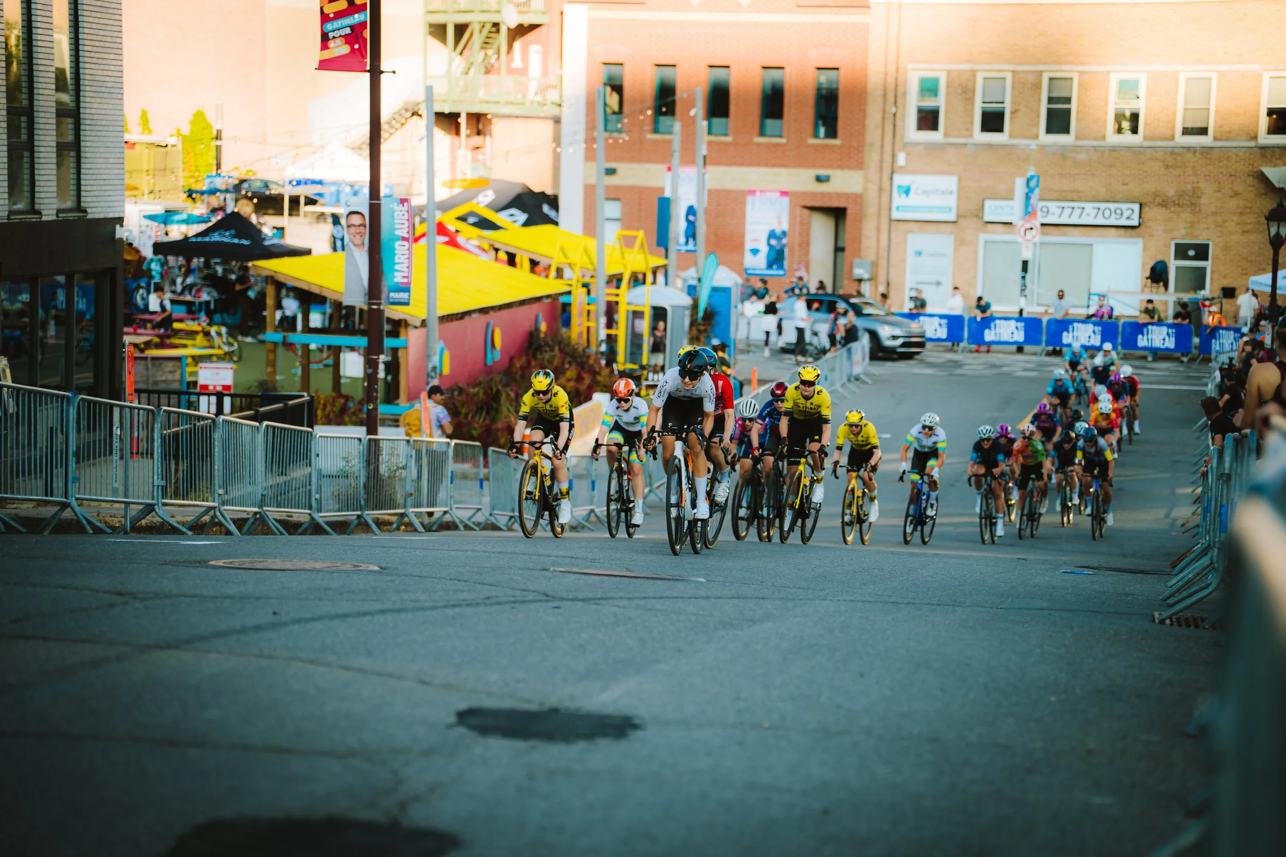 Cyclists participating in a race on a city street, with barriers and spectators on the sides, and colorful buildings and tents in the background during daylight.