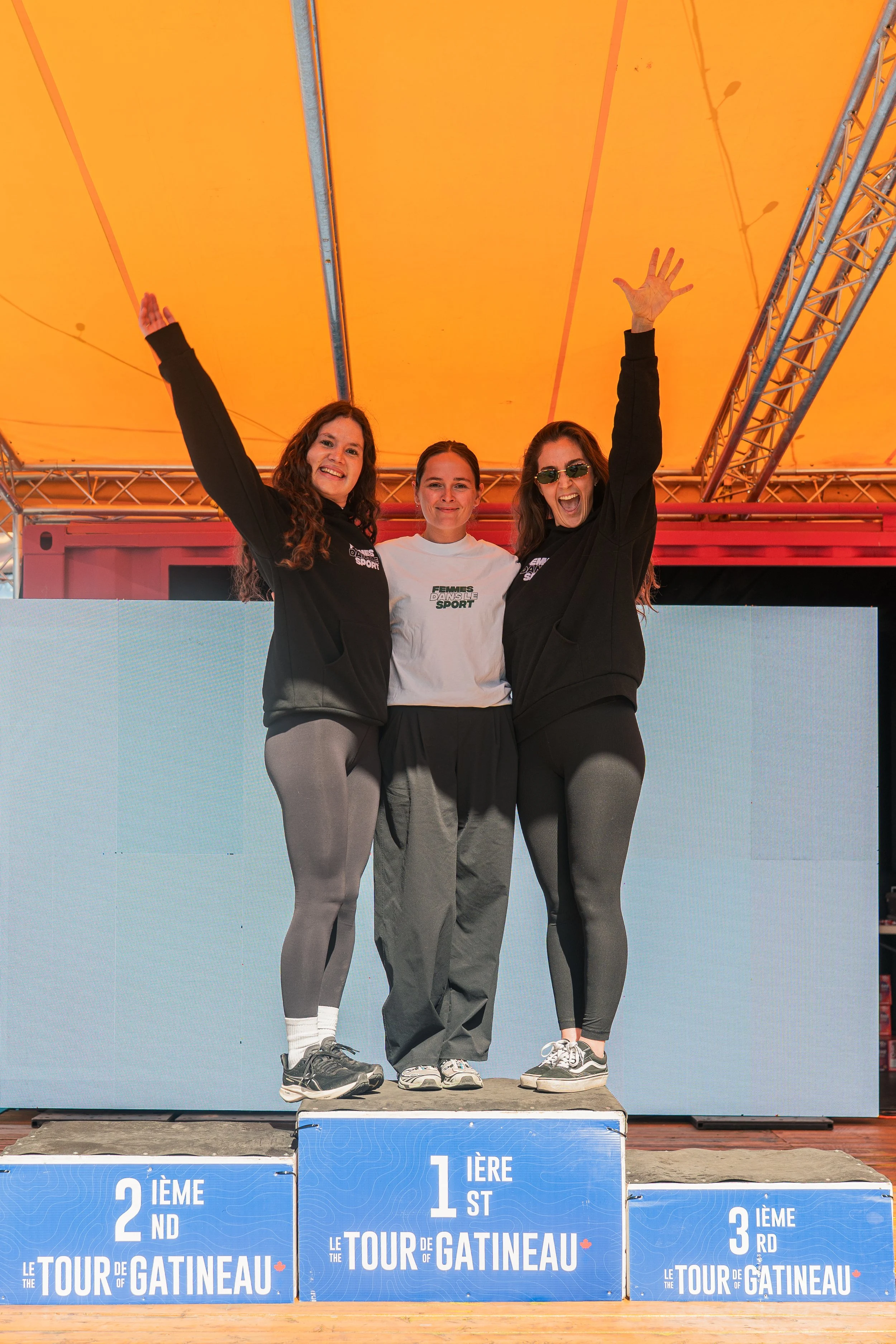 Three women standing on a winners' podium at the Tour de Gatinau event, celebrating, with the first-place winner in the middle and the second and third-place winners on the sides, under an orange canopy.