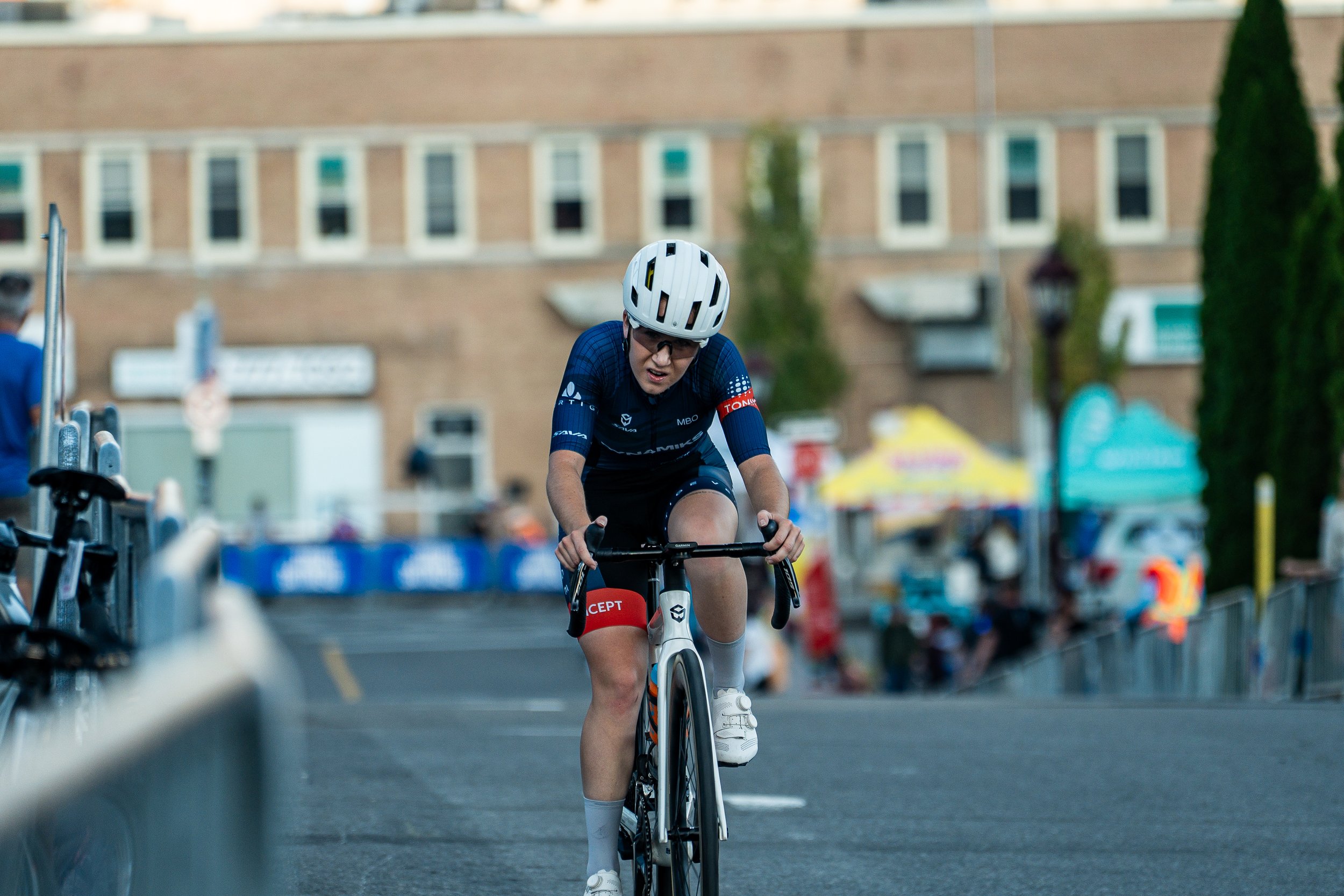 A cyclist in a race, wearing a blue and black jersey, white helmet, black shorts, and white shoes, riding on asphalt with blurred spectators, tents, and buildings in the background.