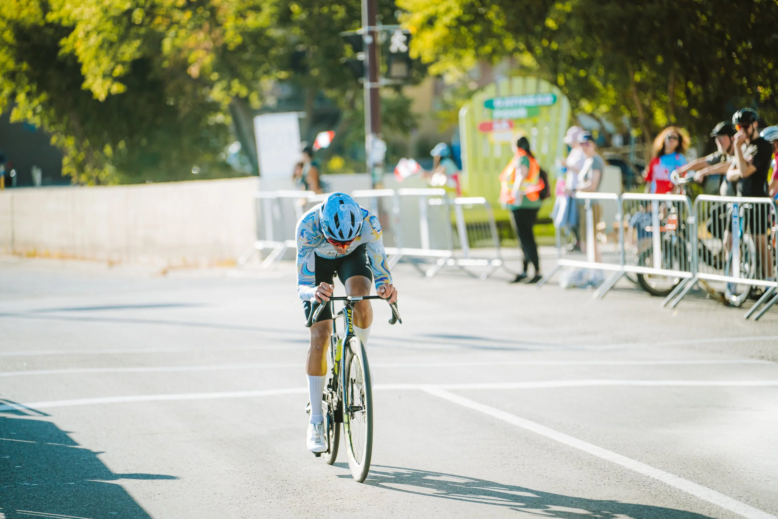 A cyclist in a blue helmet and colorful sportswear racing on a road during a competitive event, with spectators and barricades in the background.