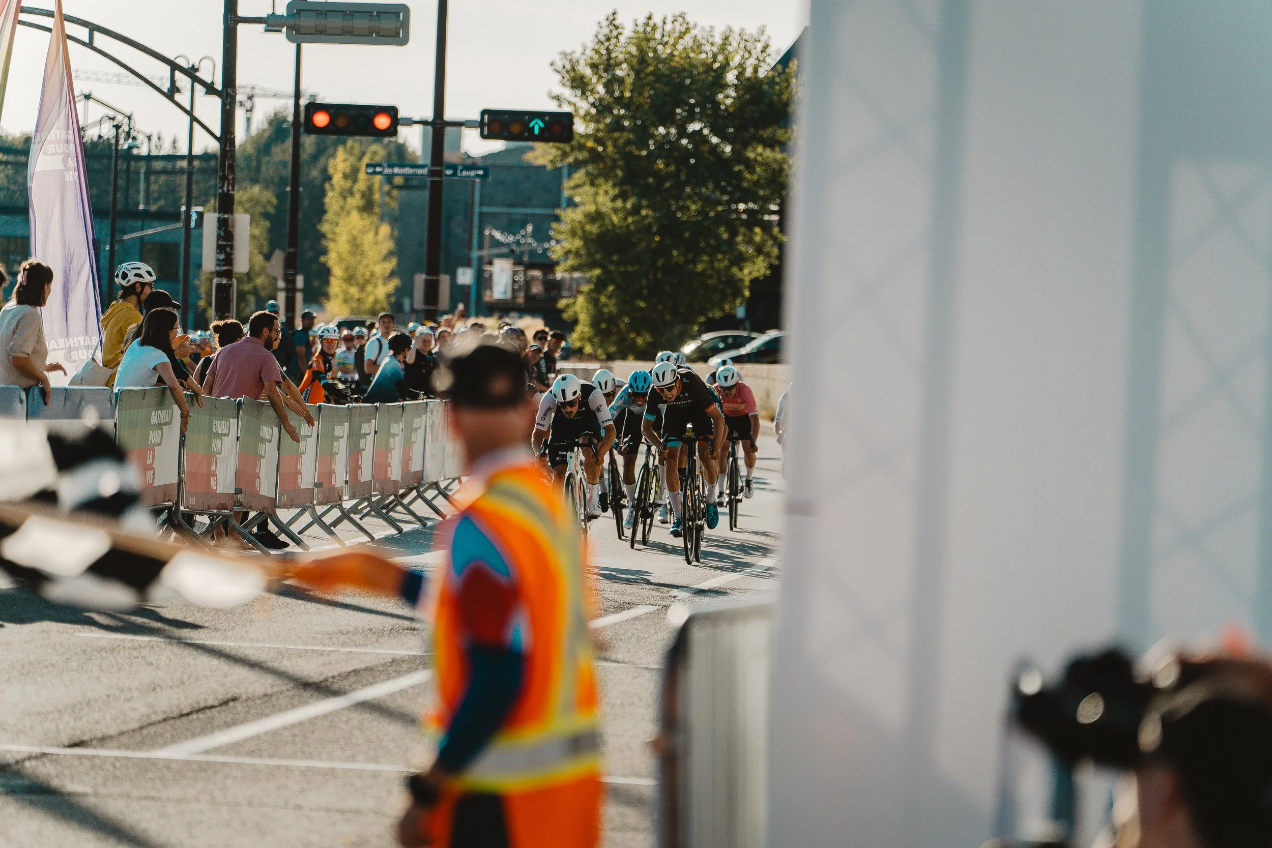 Cyclists racing in a road race with spectators watching behind barriers. The scene is outdoors on a sunny day, with some trees and traffic lights visible.