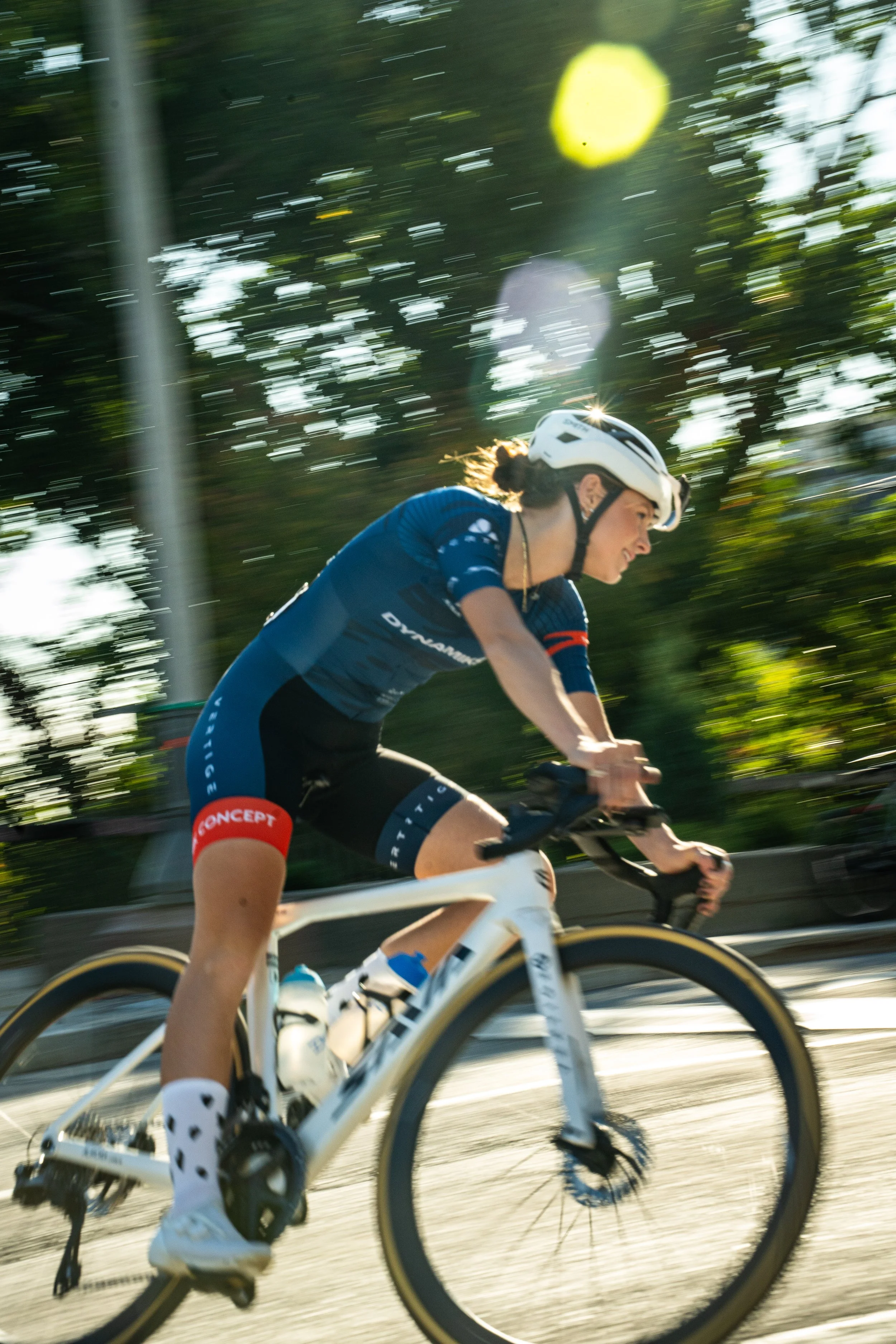 A woman cyclist riding a white bike, wearing a blue cycling kit, white helmet, and sporting polka dot socks, riding on a sunny outdoor path surrounded by trees.