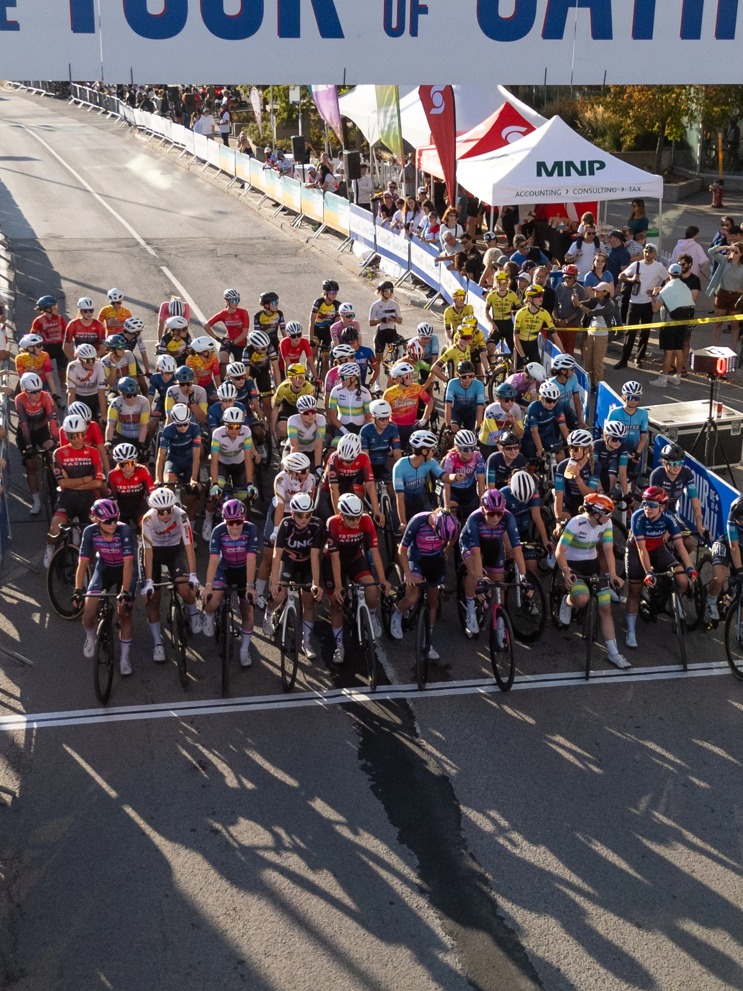 A large group of cyclists at the start line of a bike race, wearing colorful jerseys and helmets, ready to begin. Spectators and tents line the roadside behind barriers.