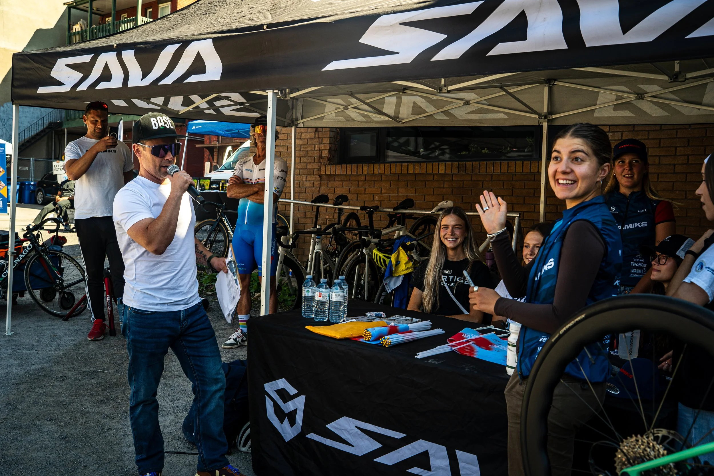 A group of people at a cycling event, with a man in sunglasses and a cap speaking into a microphone under a canopy, and smiling women sitting around a table with water bottles, posters, and event gear.