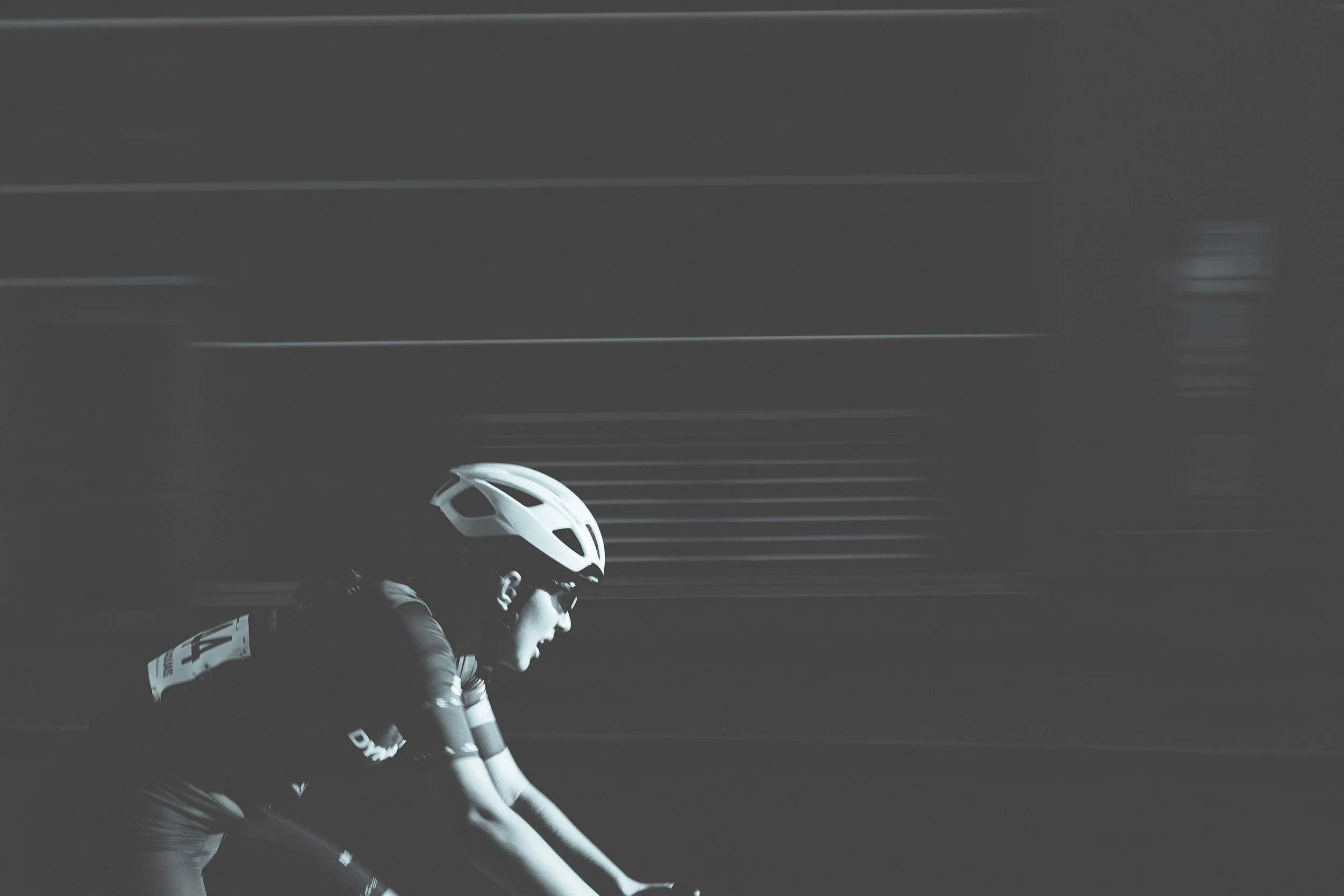 A cyclist wearing a helmet and sunglasses riding a road bike against a dark background with motion blur.