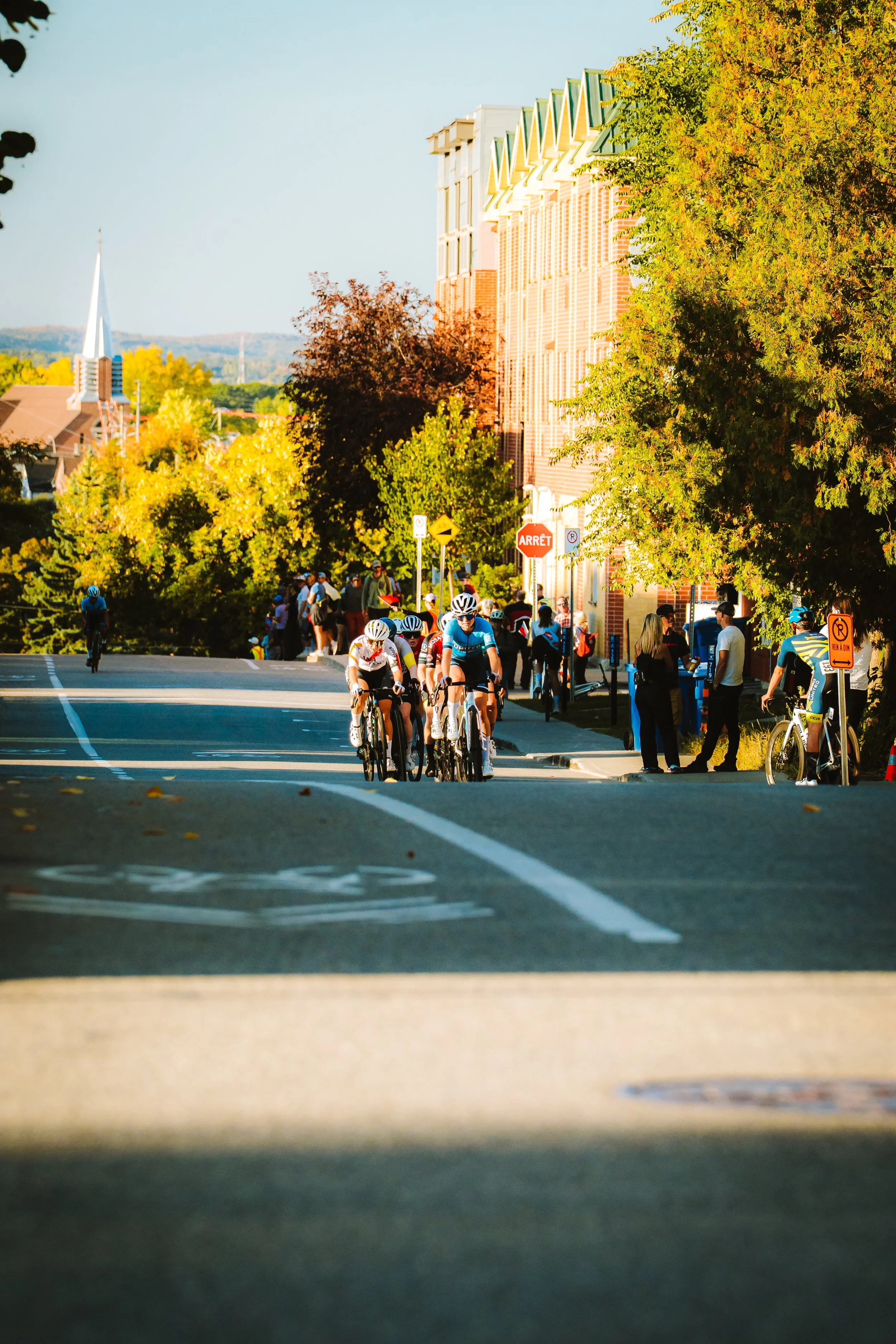 Cyclists riding on a city street during the day with trees and buildings lining the sidewalk, and people standing at the sidewalk observing.