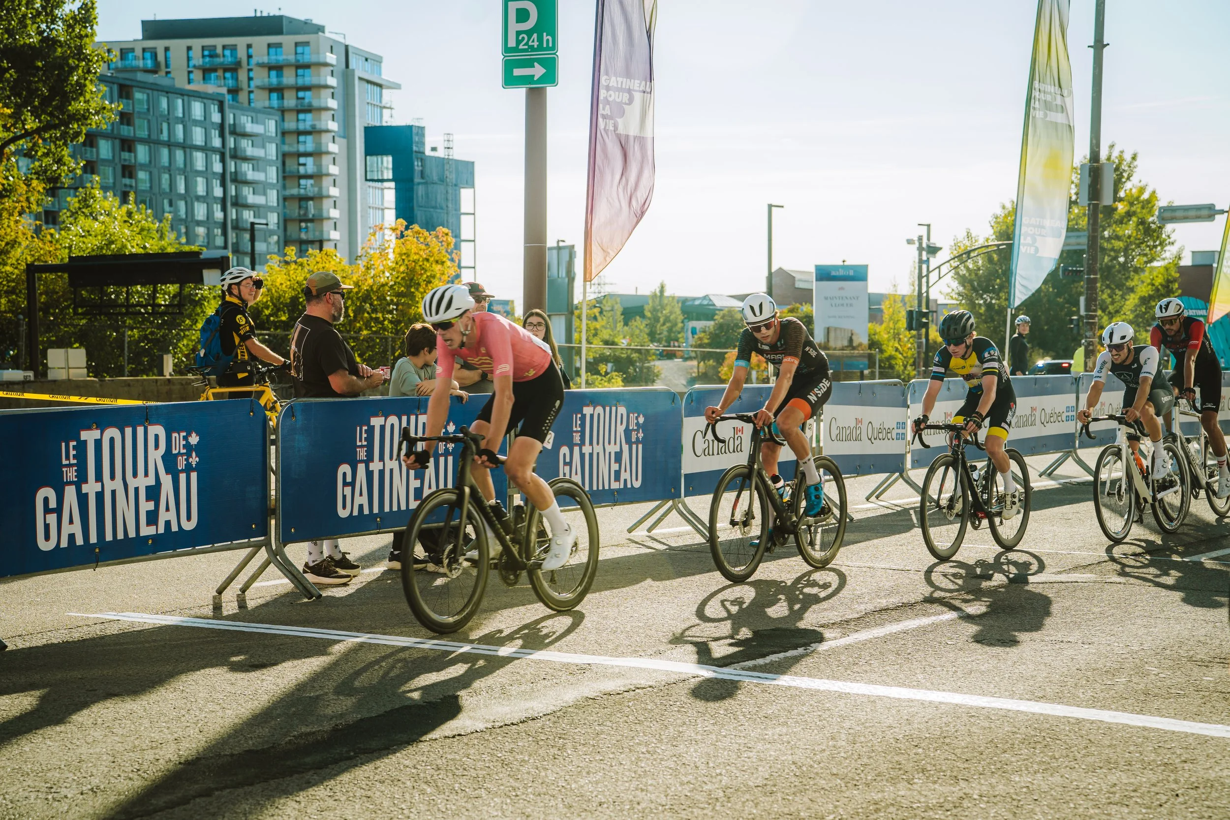 Cyclists at the start of a race during Le Tour de Gatineau cycling event in an urban area, with spectators and city buildings in the background.