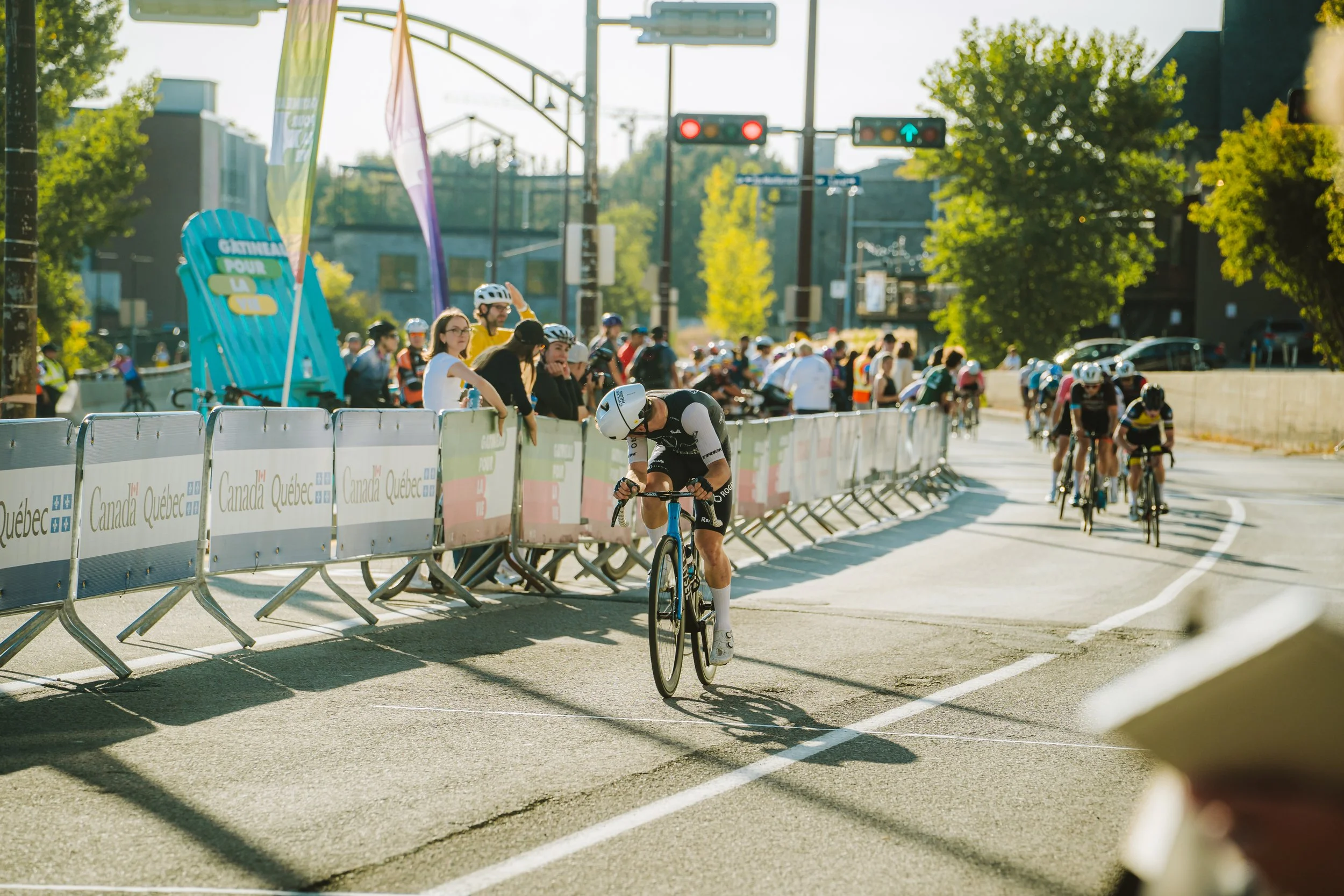 A cyclist riding with a group during a race on a city street, spectators watching behind barriers, trees, and traffic lights in the background, with sunny weather.