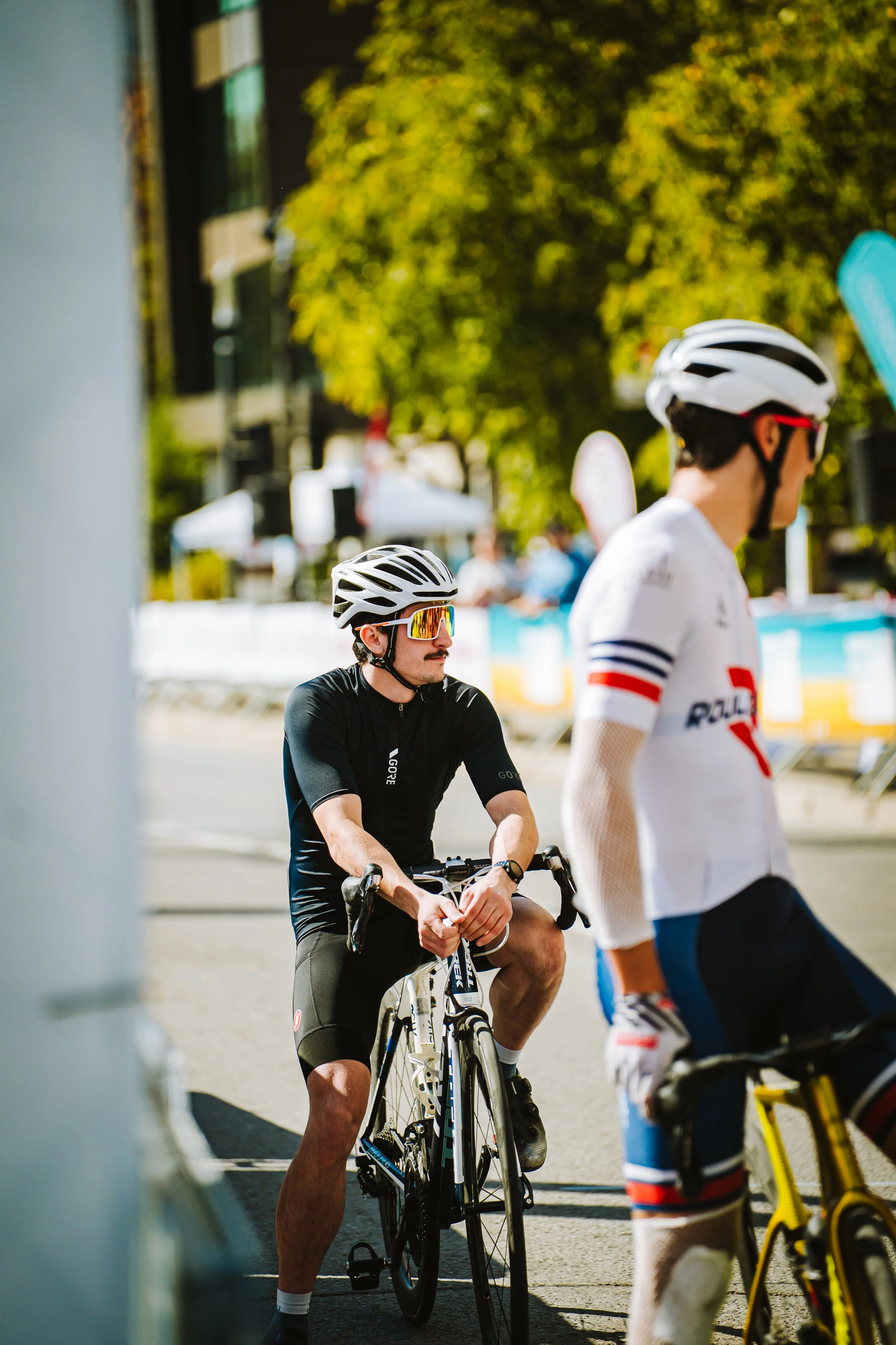 Cyclists wearing helmets and sports gear, standing with their bikes at an outdoor event on a sunny day.