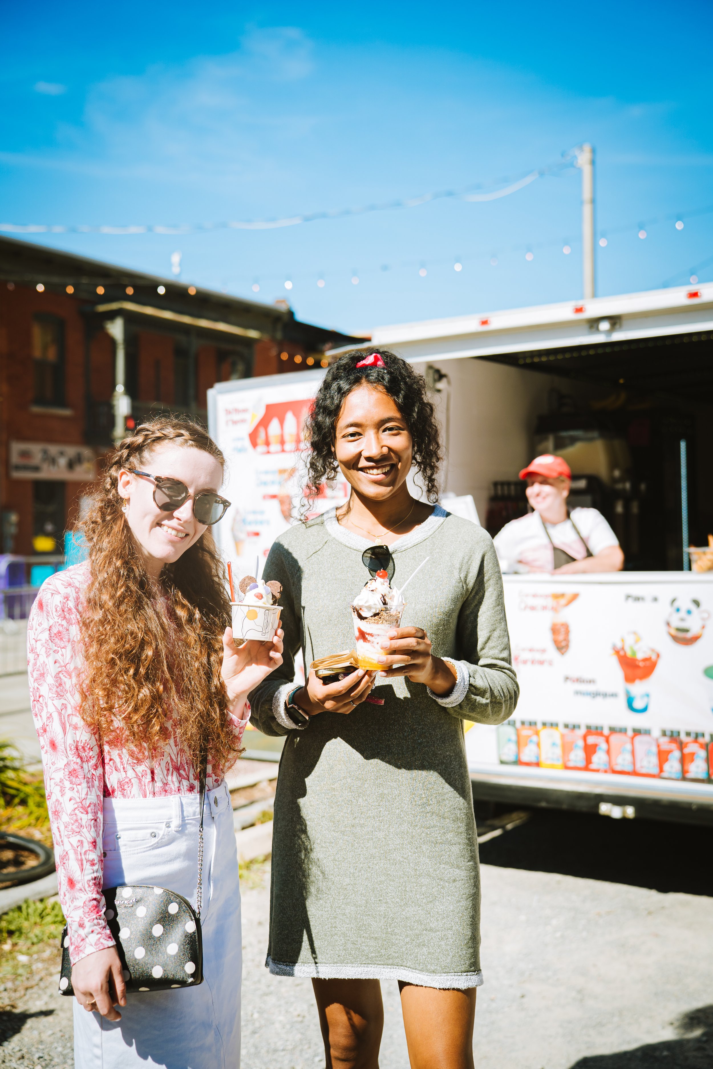 Two women standing outdoors in front of an ice cream truck, smiling and holding ice cream cones on a bright, sunny day.