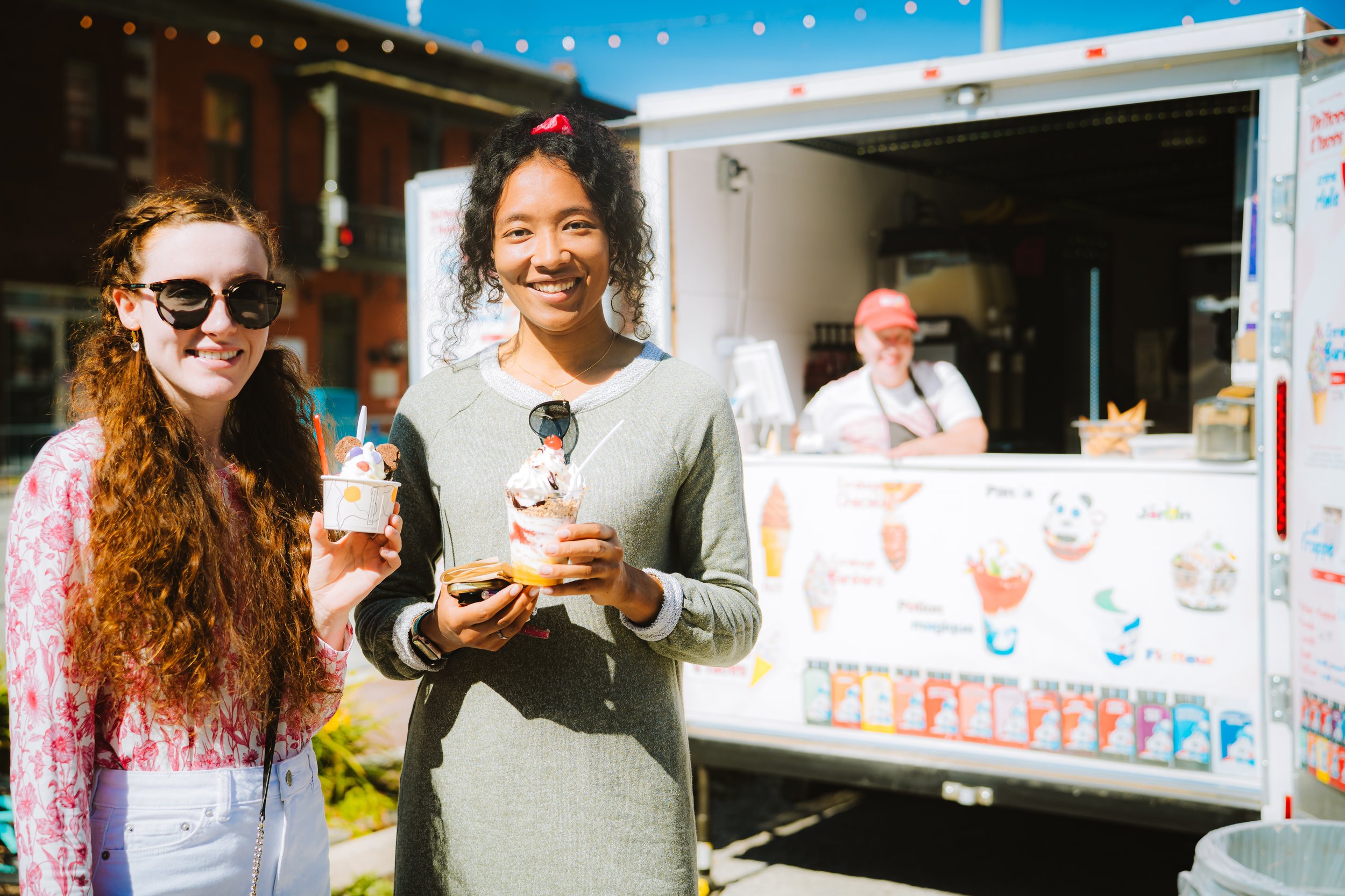Two women holding ice cream cones and enjoying ice cream outdoors on a sunny day, with an ice cream truck in the background.