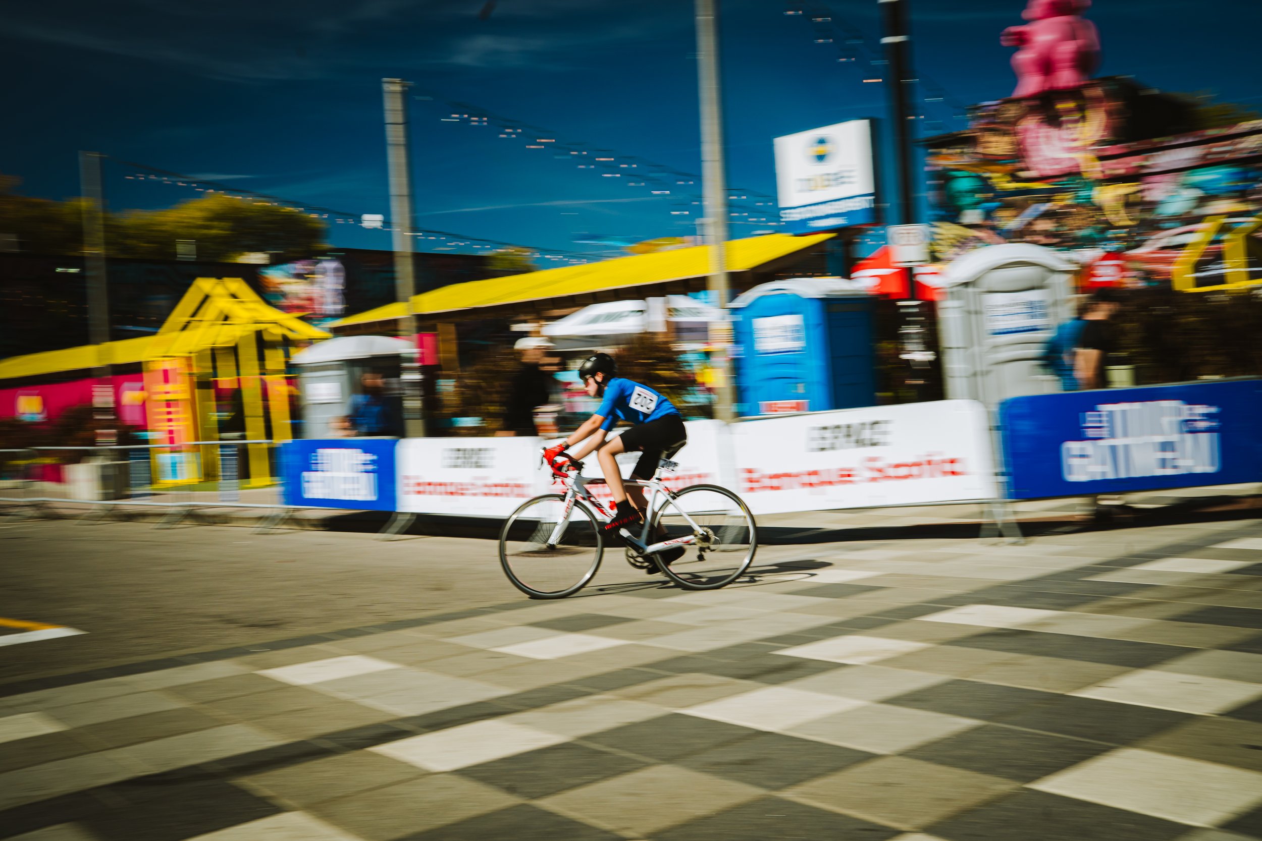A person riding a bicycle across a crosswalk at an outdoor event, with colorful carnival rides and booths in the background.