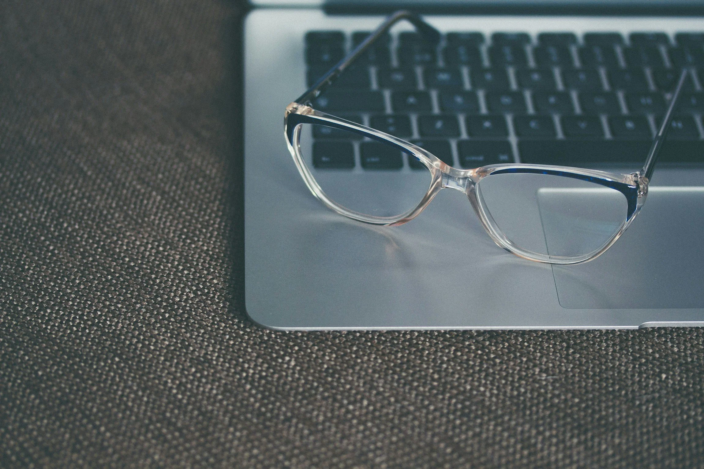 A pair of clear eyeglasses resting on a silver laptop on a dark textured surface.